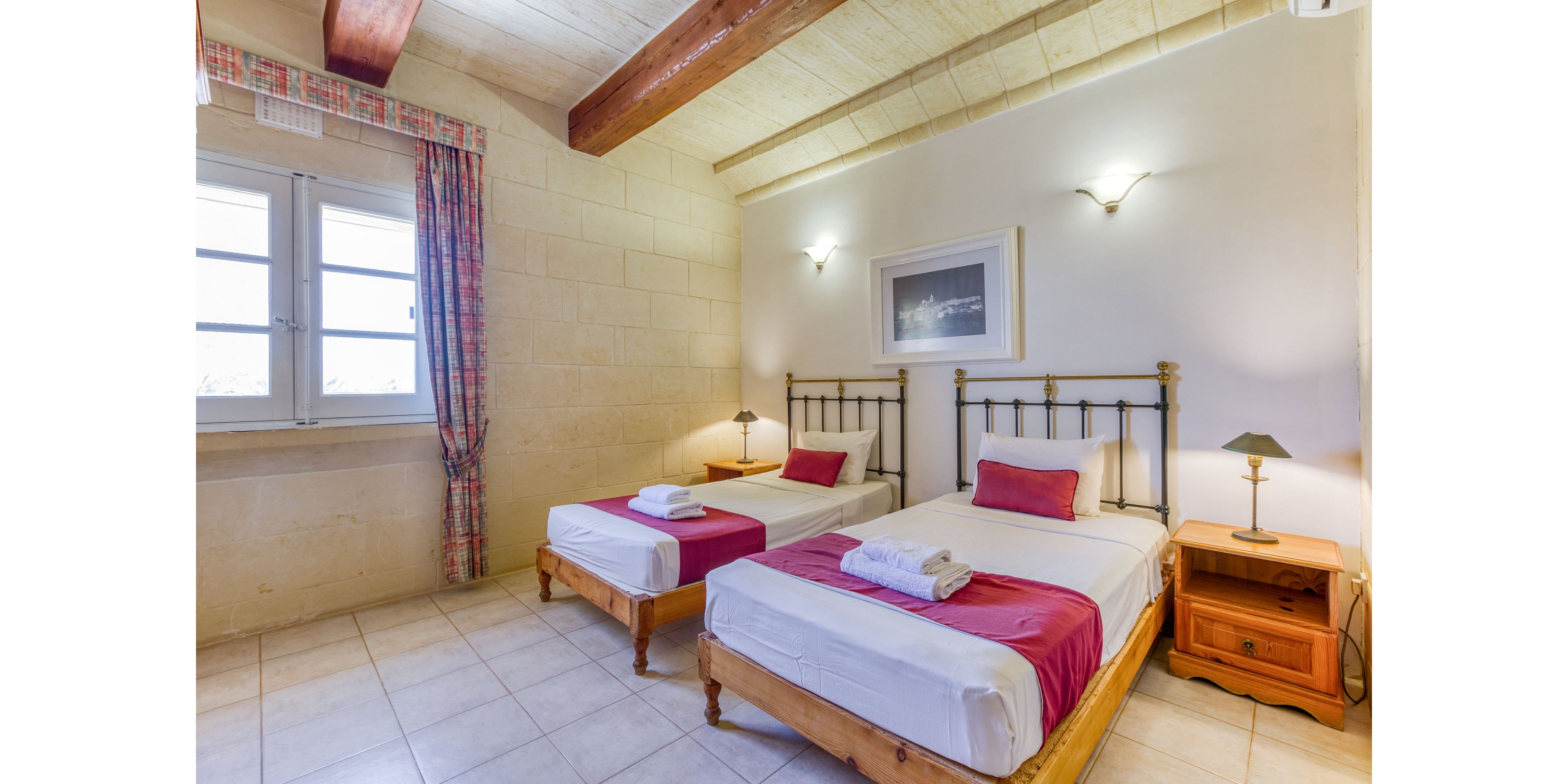 Cozy twin bedroom featuring two single beds with metal frames and wooden bases, each dressed in white linens and magenta runners. The room has light stone walls, tiled flooring, and exposed wooden ceiling beams that add a rustic touch. A window with patterned curtains lets in natural light, while wall sconces and bedside lamps provide warm illumination. A framed picture above the beds and neatly folded towels create a welcoming, hotel-like atmosphere.
