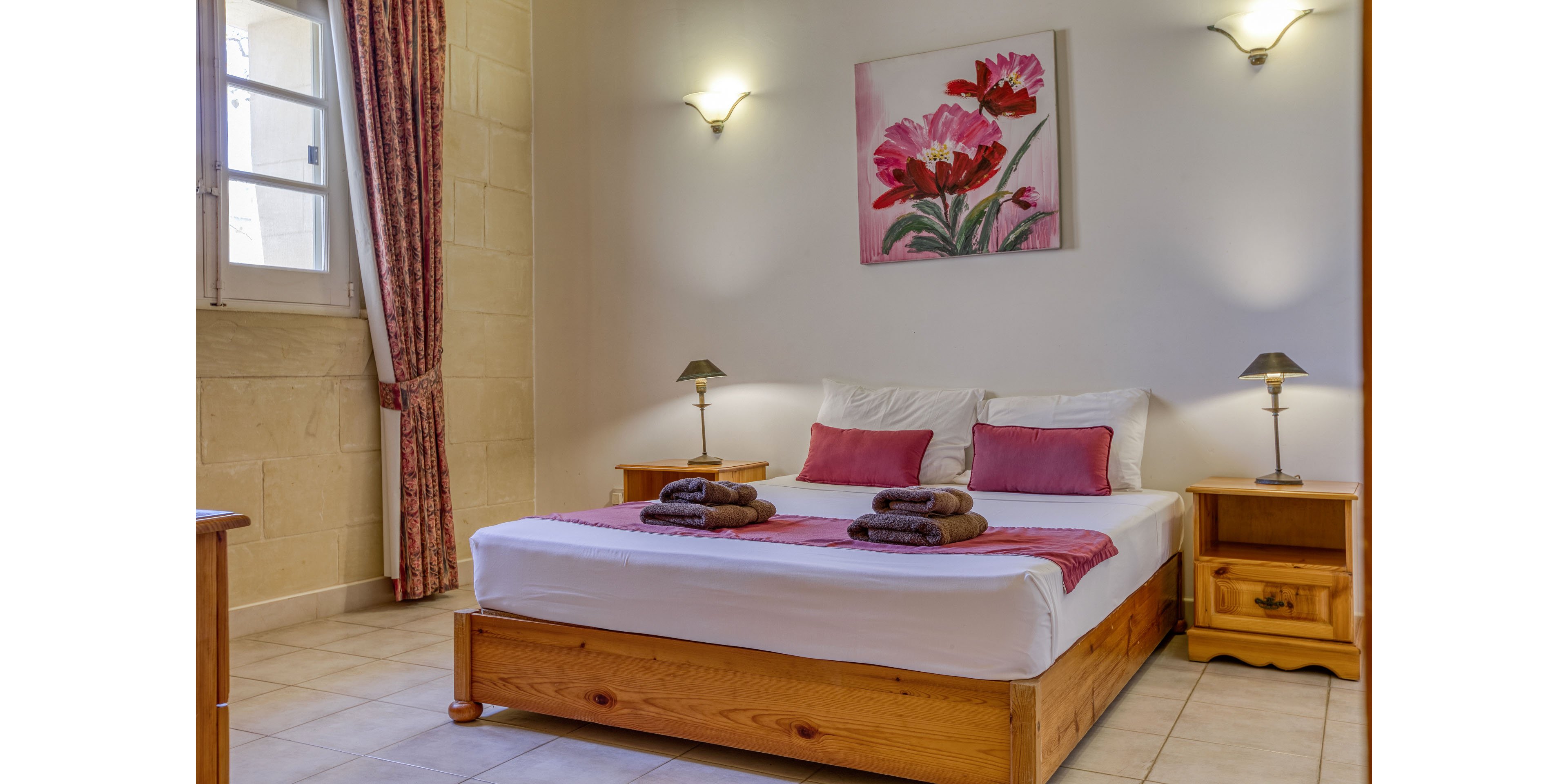 Bright, inviting bedroom featuring a wooden double bed neatly made with white linens and pink accent pillows. Rolled brown towels are placed on the bed, suggesting a guest-ready space. Matching wooden bedside tables with small lamps flank the bed, and floral artwork hangs above the headboard. A window with patterned curtains and light-colored tiled floor complete the warm, comfortable setting.