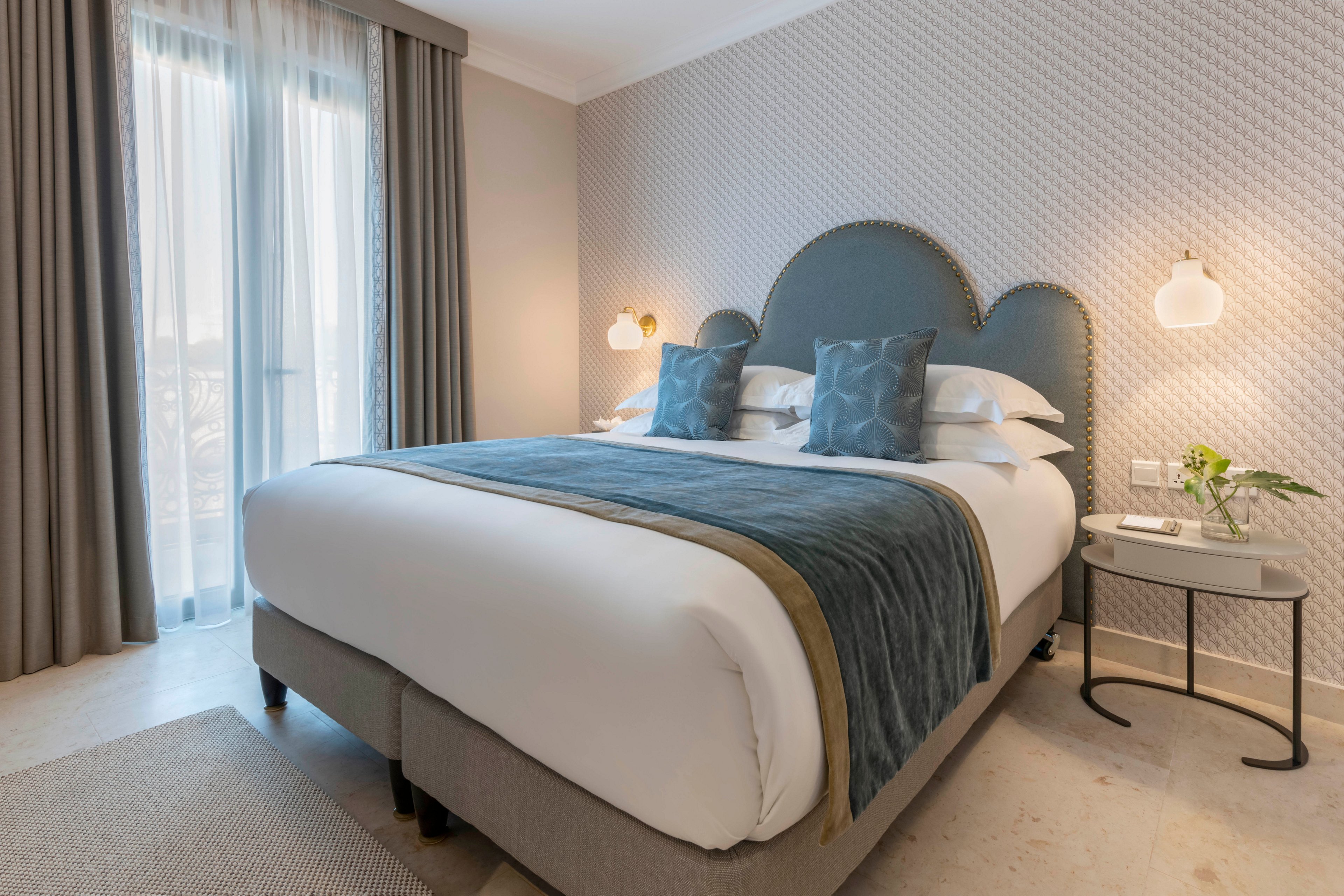 A neatly made double bed with a padded headboard is the focal point of this modern hotel bedroom. Soft natural light filters through sheer curtains covering tall windows, creating a calm and inviting atmosphere. Coordinated bedding in muted blue and gray tones, along with matching pillows and throw, enhances the contemporary style. Minimalist bedside tables, wall-mounted lamps, and a small vase with greenery complete the clean, sophisticated look.