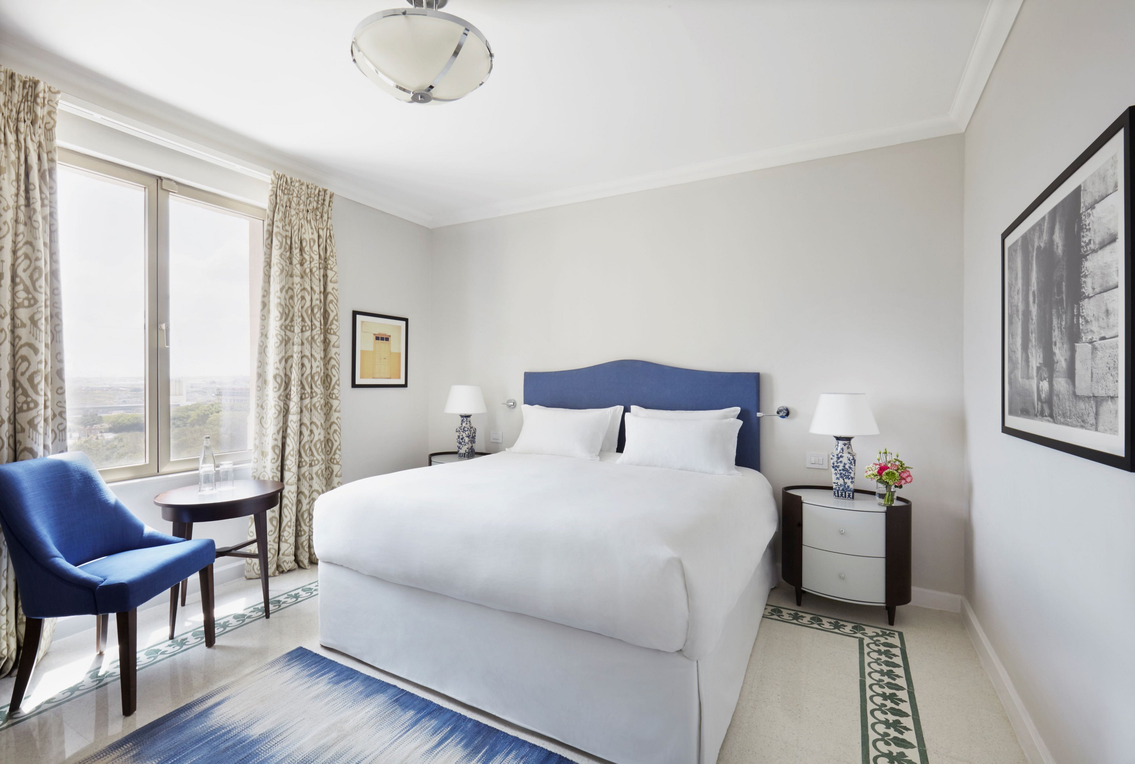 A neatly made double bed with white linens and a blue headboard is the focal point of this bright hotel bedroom. The room features a large window with patterned curtains, allowing natural light to fill the space. A blue upholstered chair and small round table sit near the window, while bedside tables with lamps and a vase of flowers flank the bed. Minimalist artwork and a blue area rug add subtle decorative touches to the clean, contemporary interior.