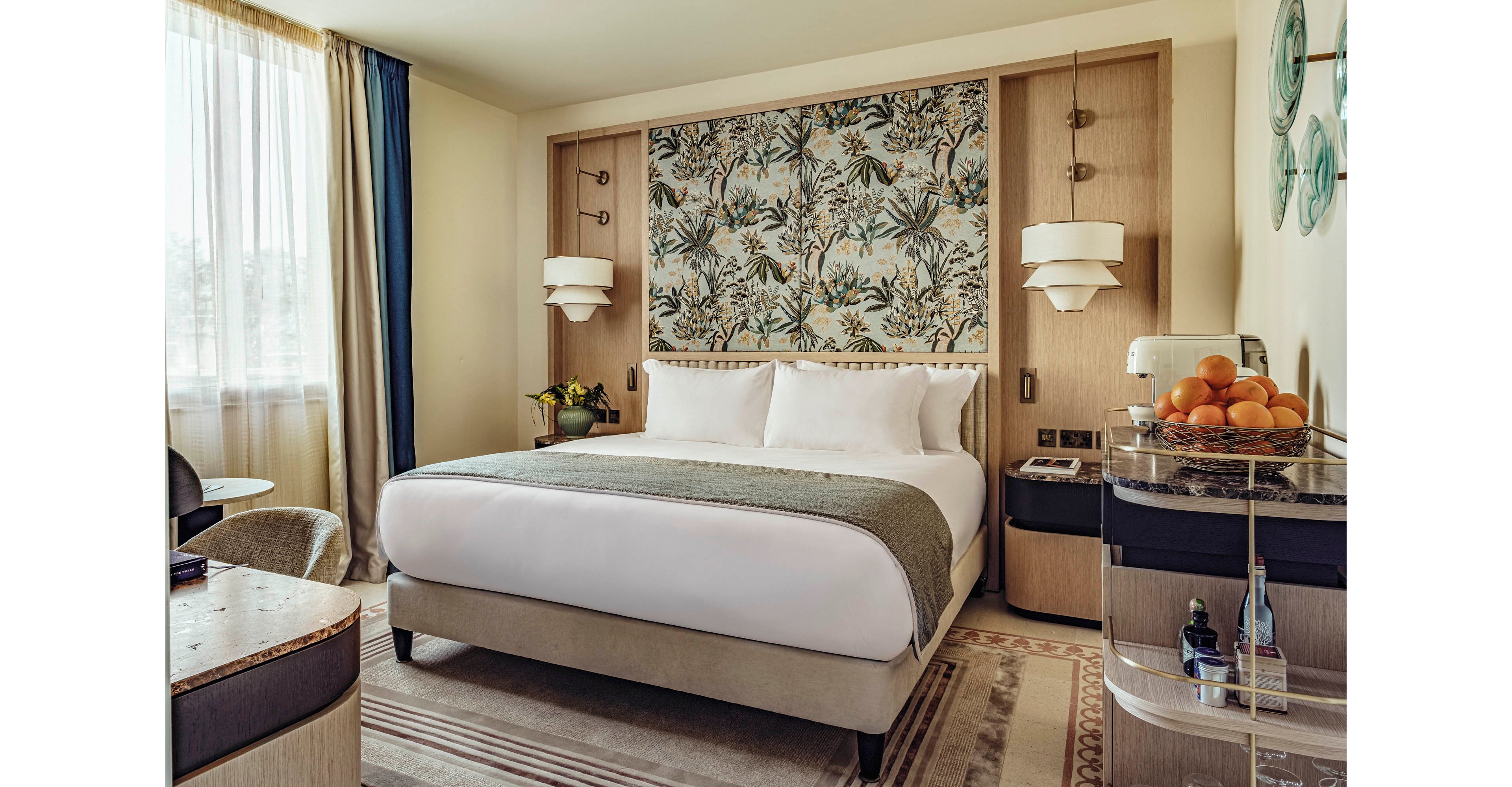 A neatly made double bed with crisp white linens stands in the center of a modern hotel room. Behind the bed, a large botanical-patterned headboard panel adds color and visual interest. Soft natural light filters through a sheer curtain beside the bed, illuminating neutral-toned furniture and decor. A side cart with a bowl of oranges and glassware adds a touch of hospitality and warmth to the space.