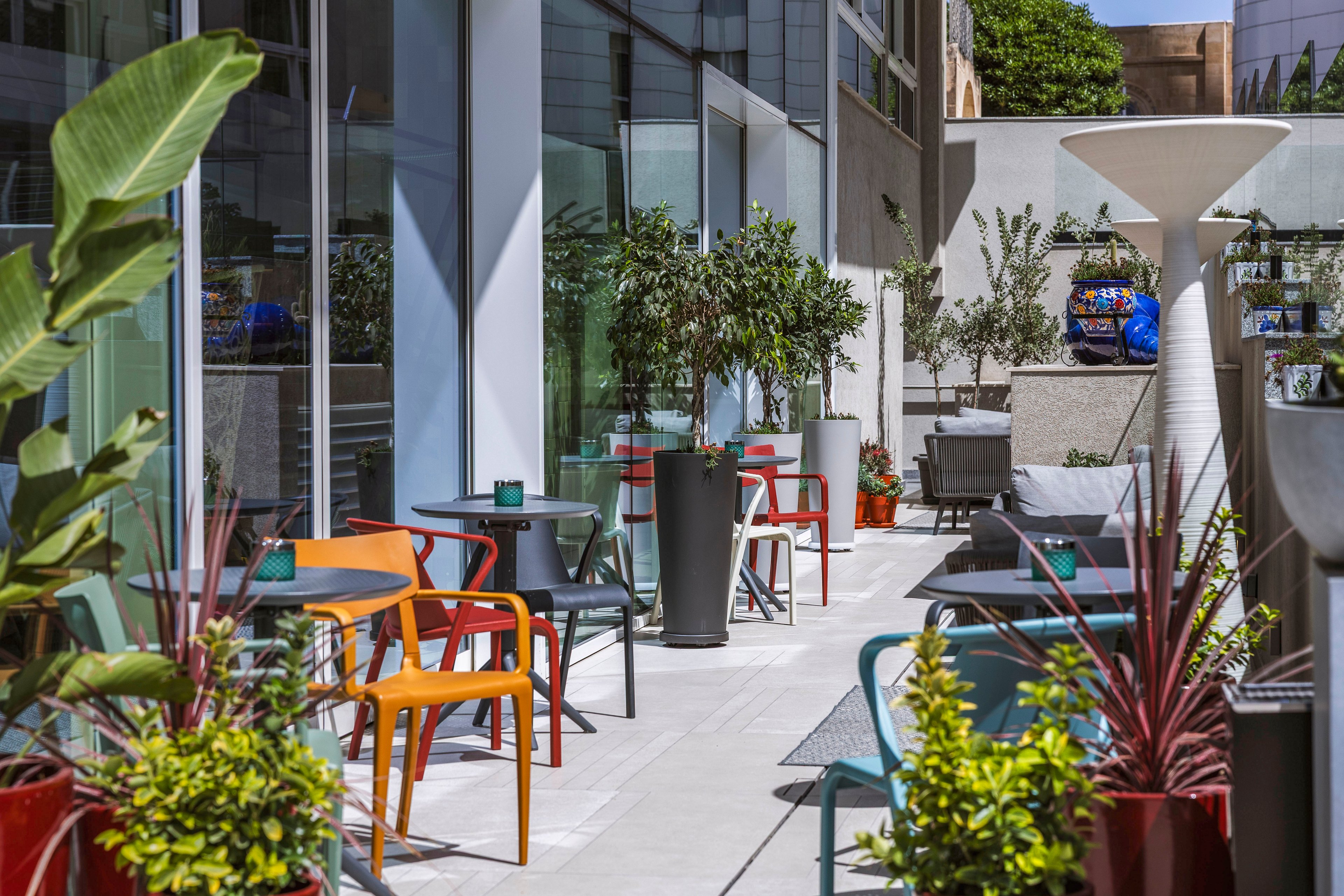 A contemporary outdoor café terrace features colorful chairs and small tables arranged along a narrow patio. Large glass windows reflect the greenery and urban surroundings, creating a bright and open atmosphere. Potted plants and shrubs in various containers line the walkway, adding lush texture and visual interest. The scene suggests a relaxed, modern setting for socializing or enjoying a drink in an urban environment.