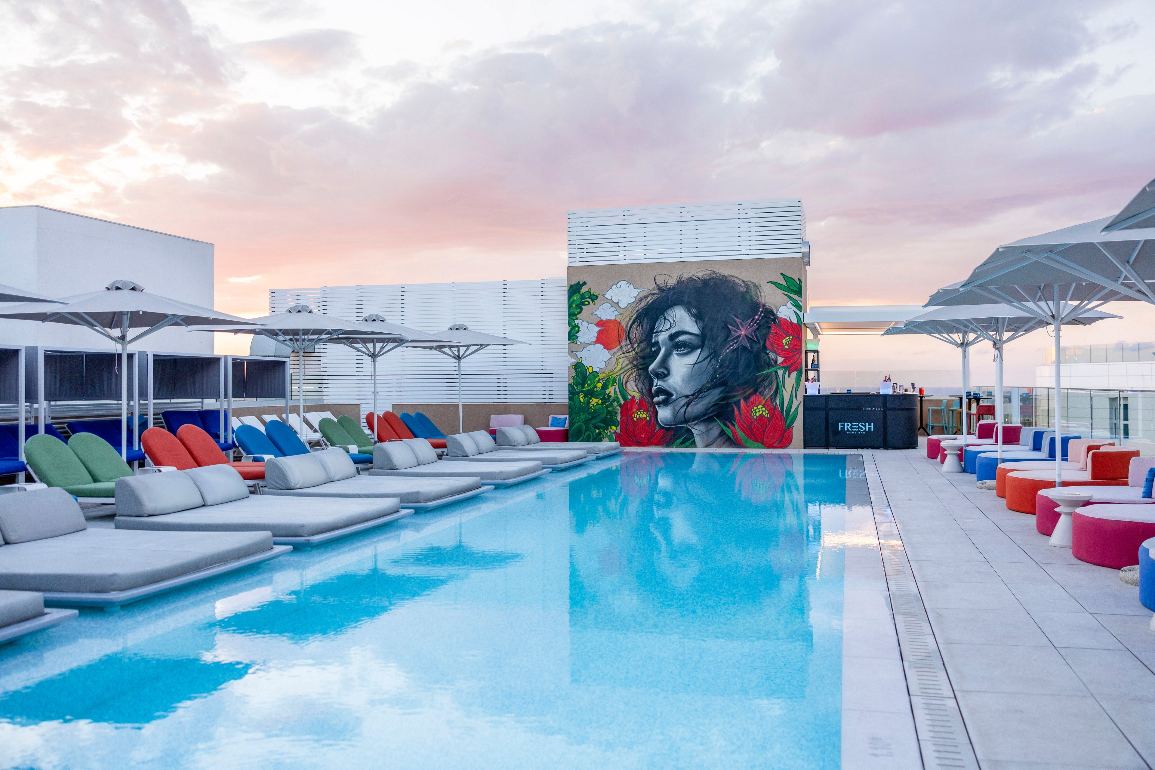 A modern rooftop swimming pool is lined with cushioned lounge beds in various bright colors. At the far end, a large mural of a person's face with floral elements adds an artistic focal point. Umbrellas provide shade along the deck, and the calm water reflects the pastel sky at sunset. The scene suggests a stylish, contemporary outdoor relaxation area in an urban setting.