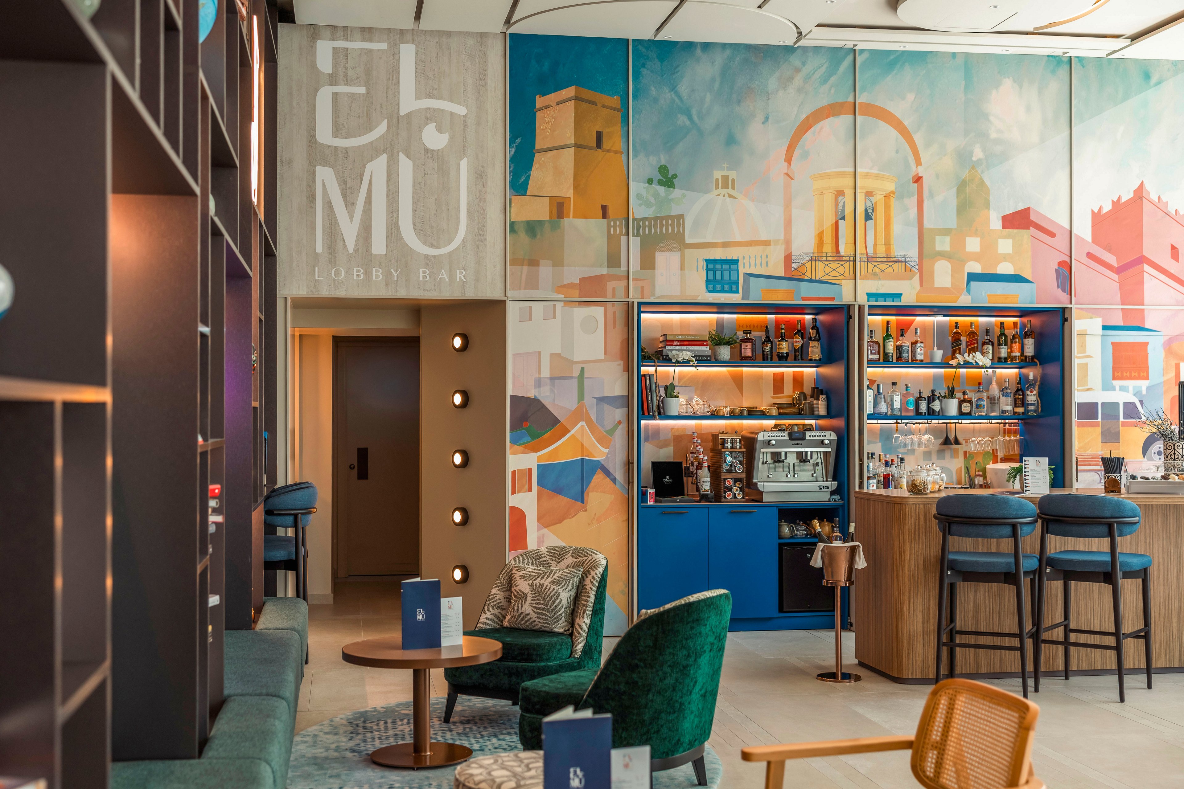 A stylish lounge bar interior features a colorful mural wall behind a well-stocked bar counter. Cozy seating with green and mustard chairs, small tables, and menus creates an inviting atmosphere. Warm lighting and contemporary decor give the space a relaxed yet sophisticated feel. The scene suggests a casual social setting suitable for drinks, coffee, or light dining.