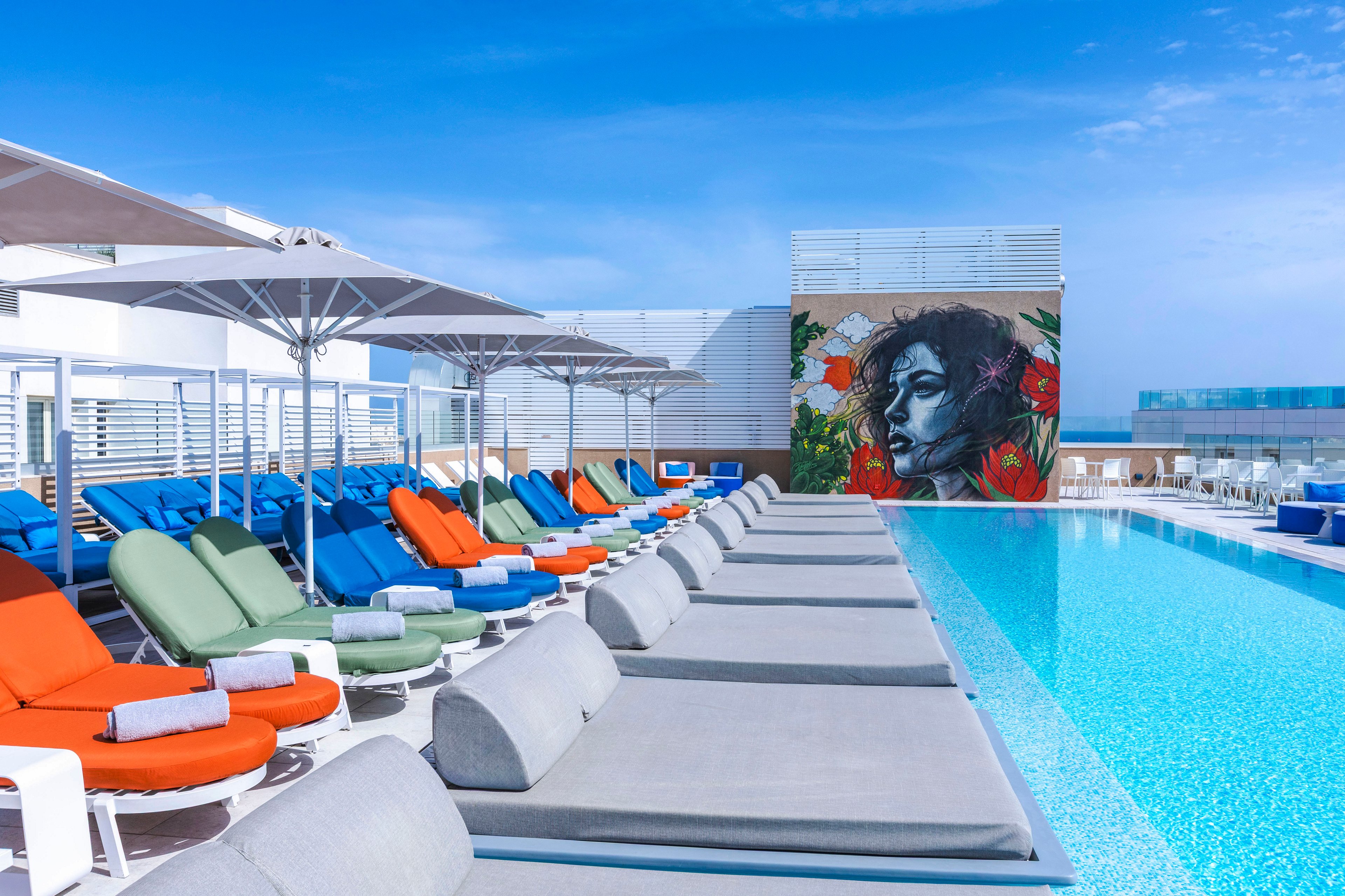 A rooftop swimming pool is lined with rows of cushioned lounge chairs in various bright colors under large white umbrellas. The clear blue water reflects the sunny sky, creating a calm and inviting atmosphere. At the far end, a large wall features a vibrant mural of a woman's face surrounded by red flowers and green leaves. The modern urban setting suggests a stylish hotel or luxury apartment terrace designed for relaxation.
