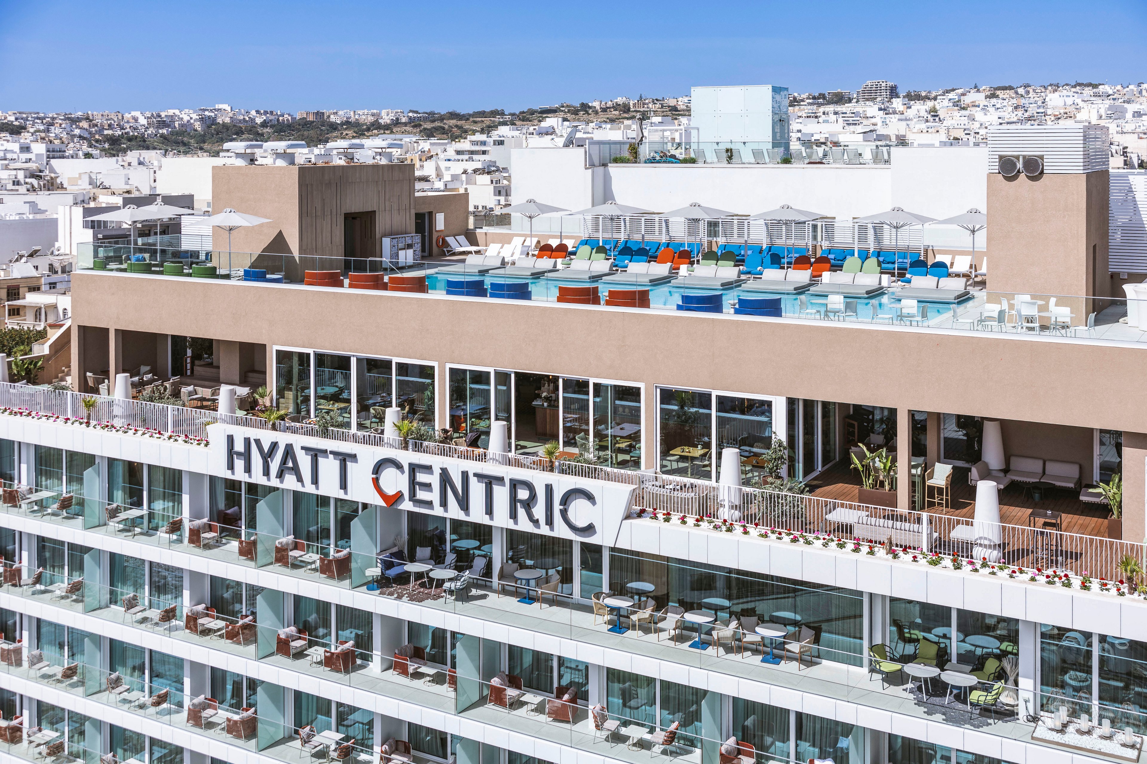 A contemporary Hyatt Centric hotel building is shown with multiple terraces and balconies overlooking a dense whitewashed cityscape. The rooftop features a swimming pool area with colorful sun loungers and umbrellas. Large glass windows reveal indoor seating and dining spaces on the upper floors. The bright daylight and clear blue sky emphasize a relaxed, upscale urban resort atmosphere.