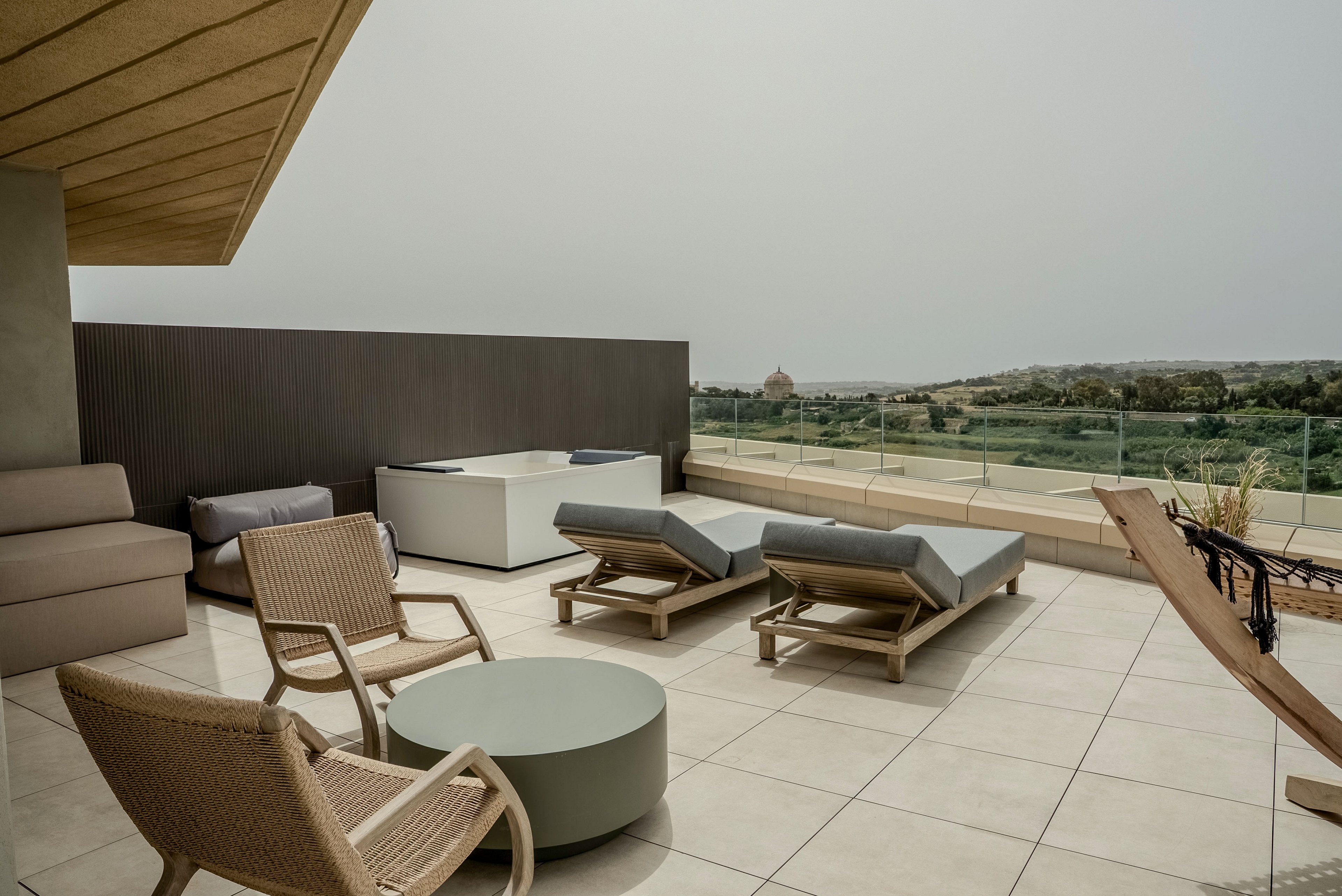 A spacious rooftop terrace features several cushioned lounge chairs and wicker armchairs arranged on light tiled flooring. A low round table and a hammock add to the relaxed outdoor seating area. Glass railings reveal a scenic view of greenery and distant buildings under an overcast sky. The neutral color palette and clean lines create a modern, minimalist atmosphere ideal for relaxation.