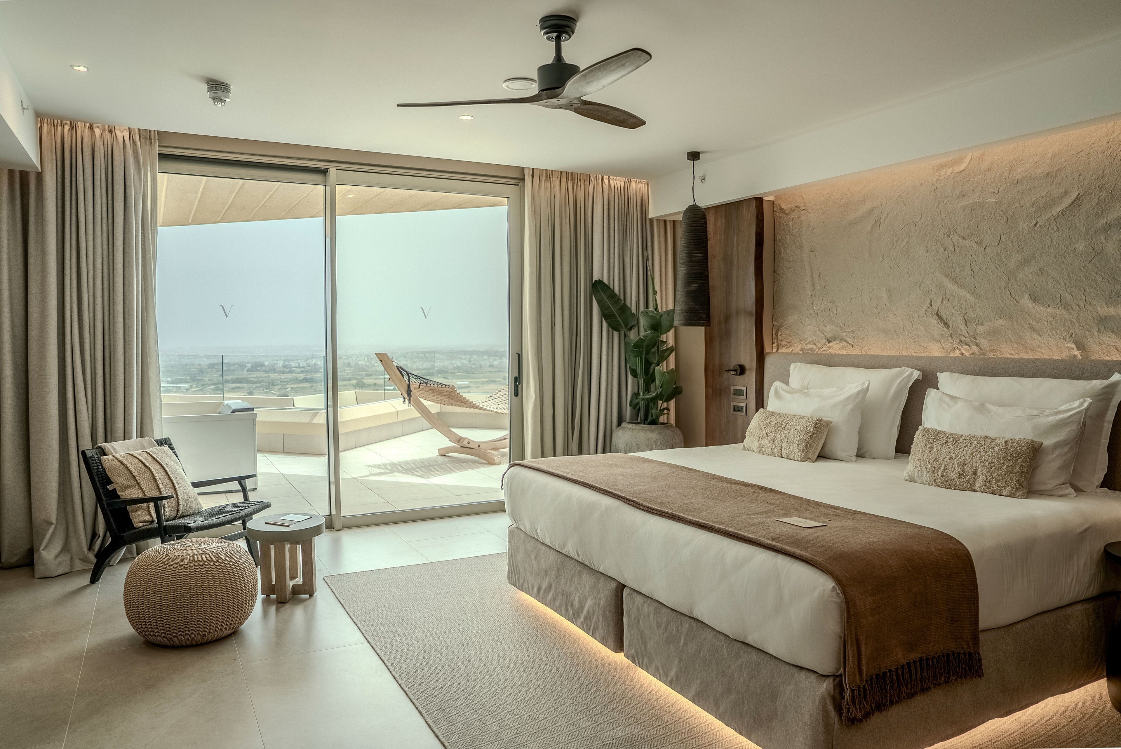 A contemporary hotel bedroom features a large double bed with neutral linens and brown accents. Floor-to-ceiling glass doors open onto a terrace with lounge chairs overlooking a distant coastal landscape. Soft lighting under the bed frame and along the ceiling enhances the warm, minimalist decor. A ceiling fan, indoor plant, and simple seating area contribute to the relaxed, resort-like atmosphere.