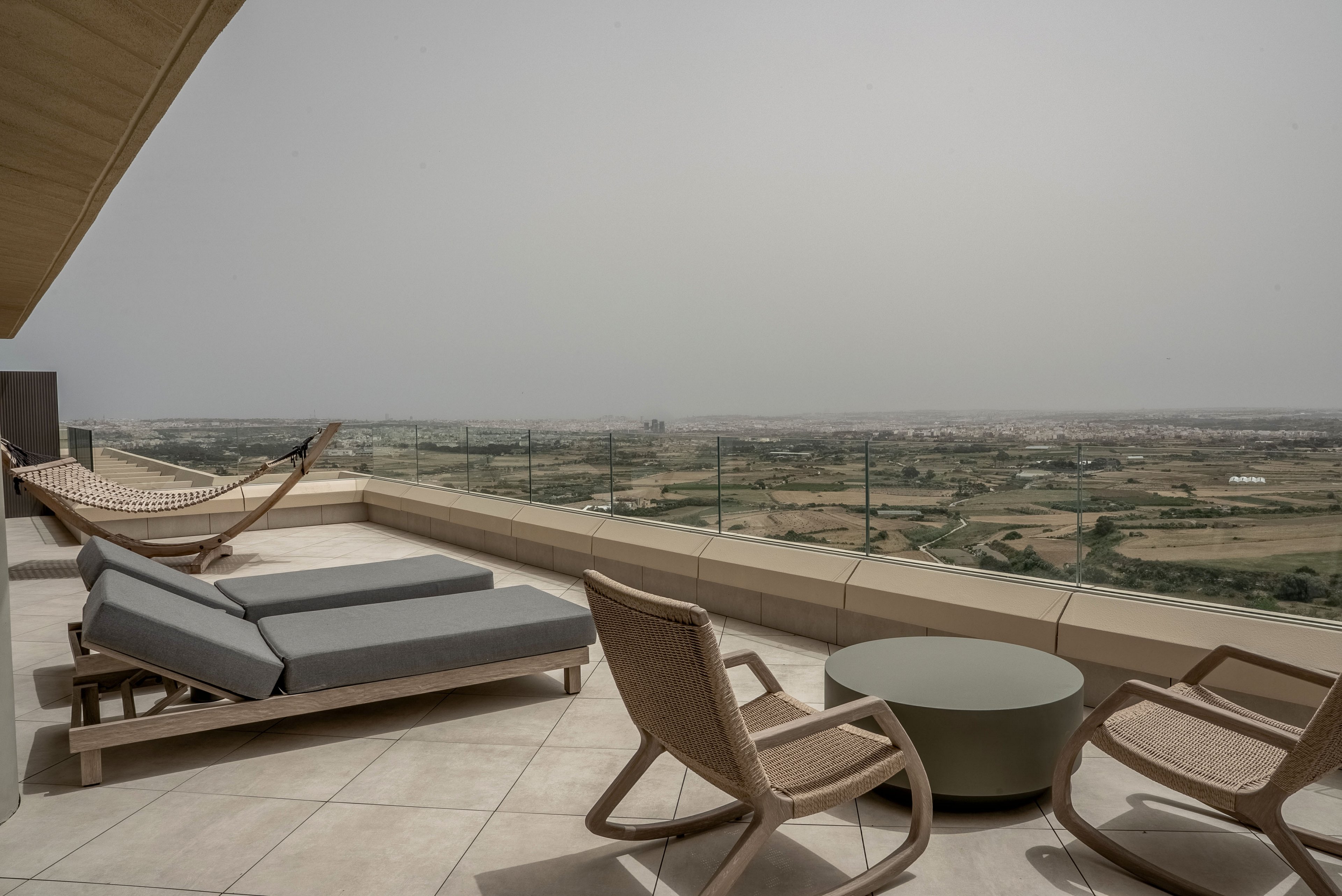 A spacious rooftop terrace features contemporary lounge chairs and a hammock arranged on light tiled flooring. Glass railing lines the edge, opening to a wide view of distant fields and low-rise buildings. Neutral tones and minimalist furniture create a calm, modern atmosphere. The hazy sky adds to the relaxed, tranquil mood of the outdoor space.