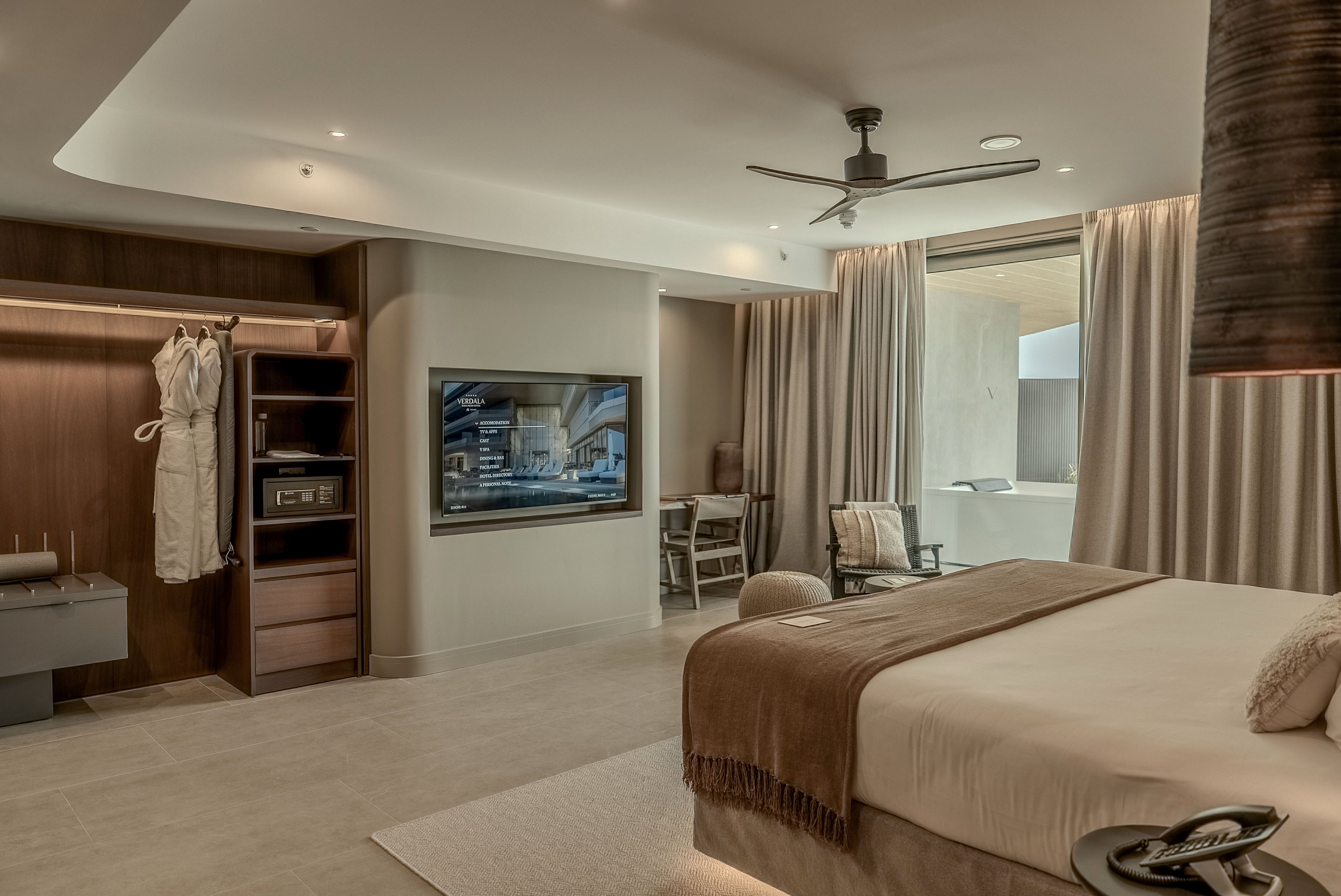 A contemporary hotel bedroom features a large bed with neutral-toned linens and a throw blanket. An open wardrobe with hanging robe, shelves, and a safe sits opposite a wall-mounted flat-screen television. Floor-to-ceiling curtains frame a doorway leading to a balcony or terrace with outdoor seating. Soft, warm lighting and minimalist decor create a calm, upscale atmosphere. The overall palette is beige, brown, and cream with sleek modern finishes.