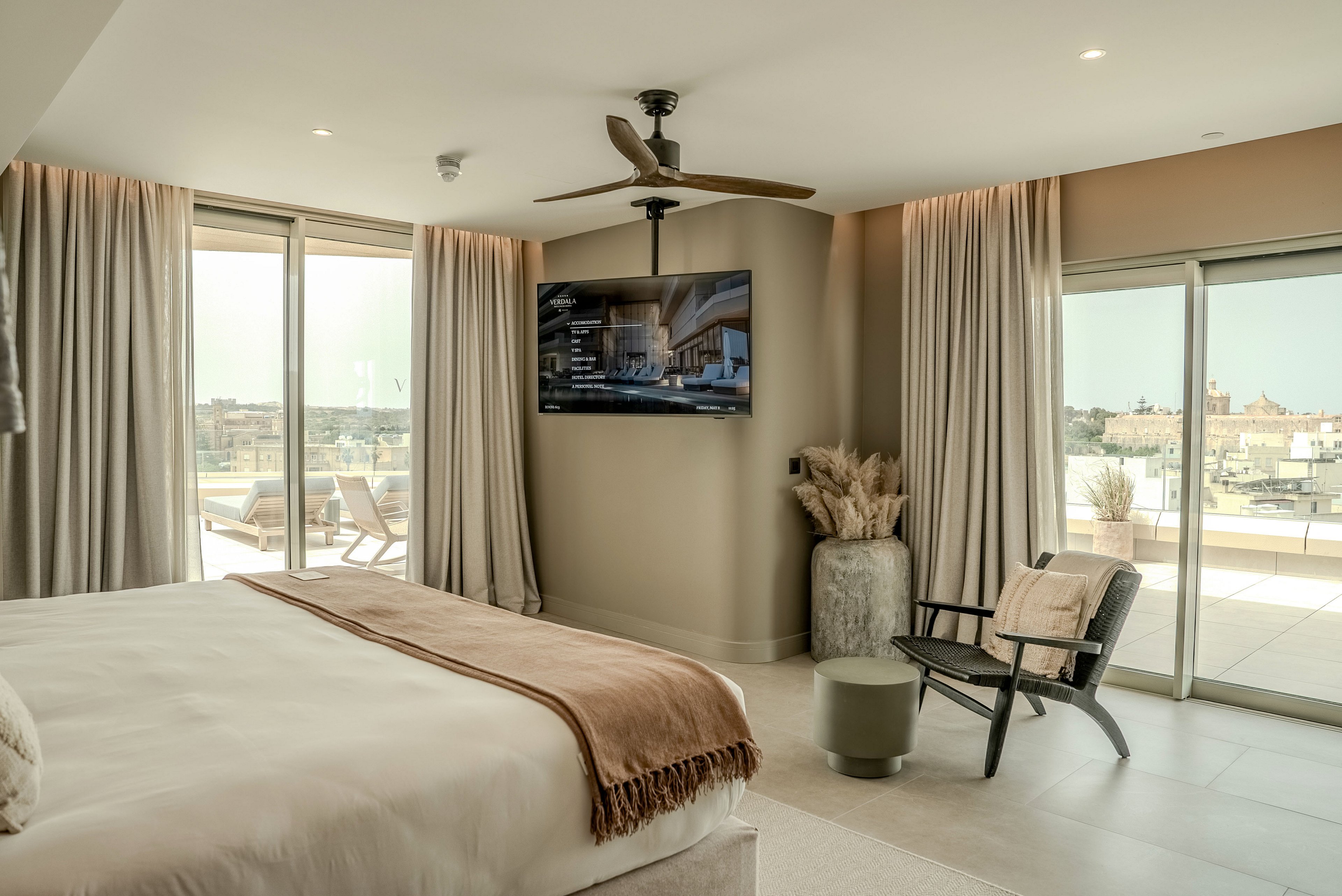 A contemporary hotel bedroom features a large bed with neutral linens and a soft throw blanket. Floor-to-ceiling curtains frame expansive sliding glass doors that open onto a sunny terrace with outdoor seating. A wall-mounted flat-screen television faces the bed, while a ceiling fan hangs above. A lounge chair, side table, and decorative vase with dried grasses add to the minimalist, beige-toned decor.