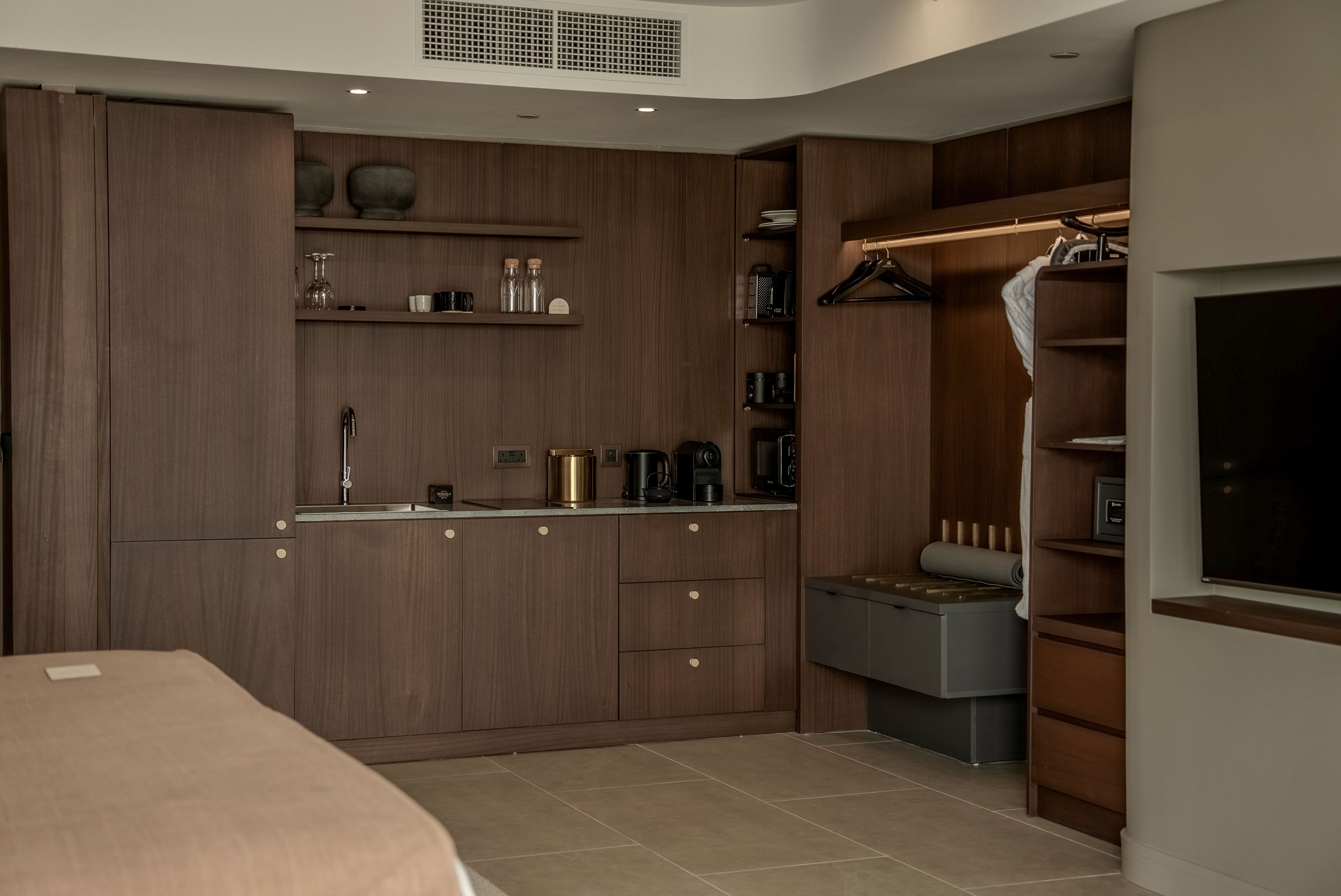 A contemporary hotel room features a compact kitchenette built into warm wooden cabinetry. The space includes a small sink, countertop appliances, and open shelves with neatly arranged dishes and bottles. An adjacent open wardrobe holds hangers and folded linens, adding to the room’s functional design. Neutral tones and soft lighting create a calm, minimalist atmosphere.