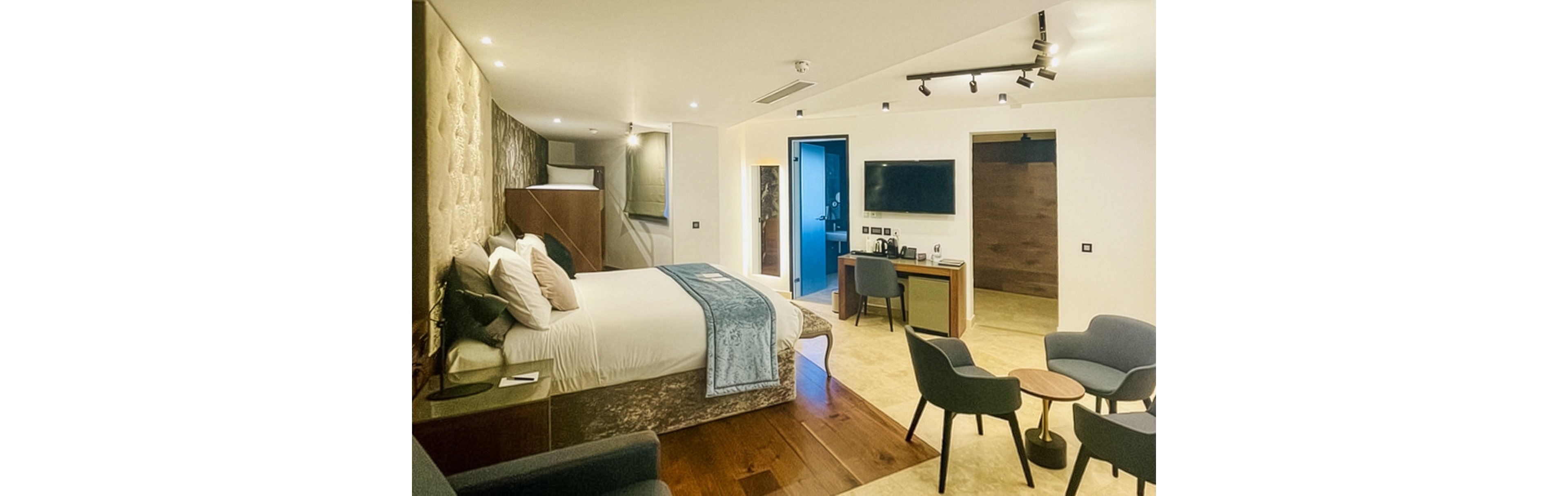 Spacious contemporary hotel room featuring a large bed with plush pillows and a soft blue throw. The suite includes a small seating area with armchairs and round tables, facing a wall-mounted television and desk. Warm recessed lighting and wood flooring create a comfortable, inviting atmosphere. Neutral tones with blue accents give the space a modern yet relaxing feel.