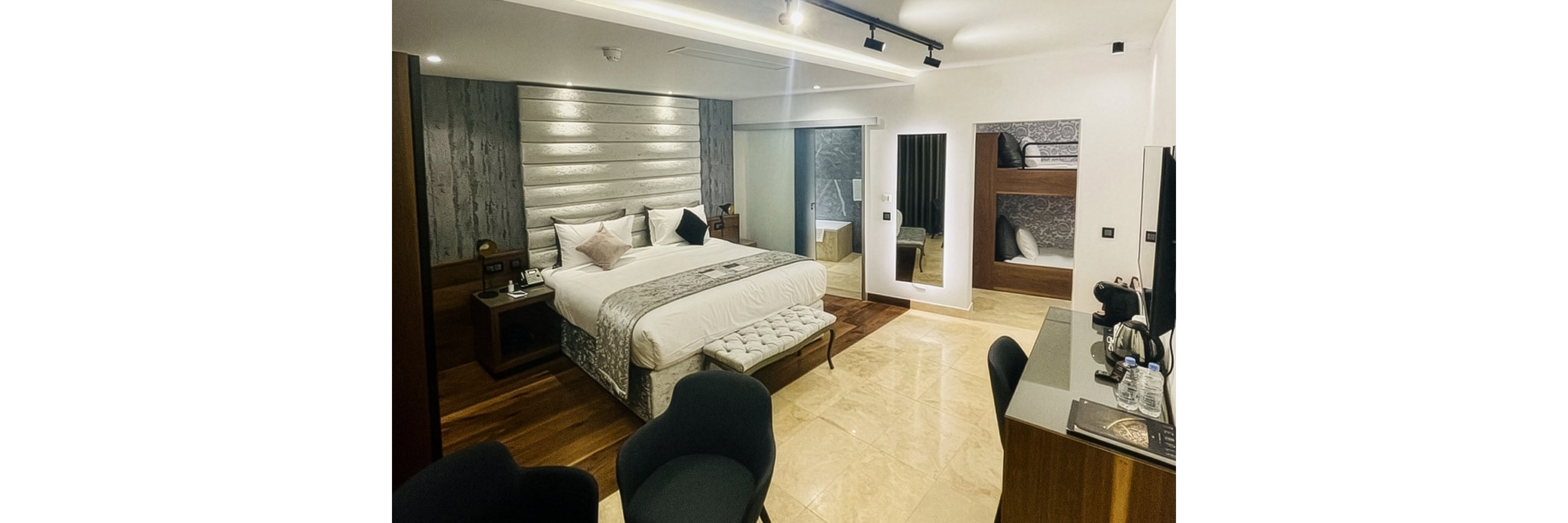 Contemporary hotel room featuring a large double bed with decorative pillows and a cushioned bench at the foot. The space includes a sleek work desk with chair, bottled water, and a coffee station. A wall-mounted mirror and recessed lighting enhance the bright, neutral interior with wood and tile flooring. Built-in bunk beds and a glass partition leading to the bathroom add functionality and style.