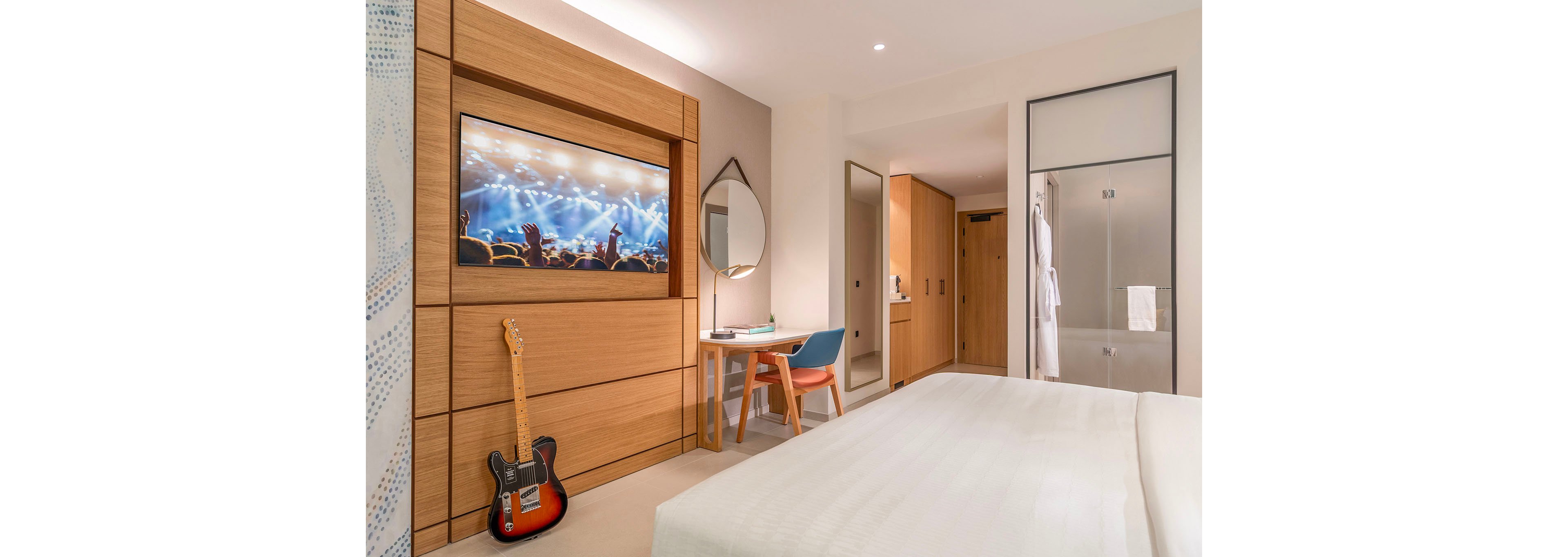 A contemporary hotel room features a large bed facing a wall-mounted television showing a concert scene with bright stage lights and cheering crowd. A wooden desk with a round mirror and colorful chair sits beside the TV unit, adding a workspace to the room. An electric guitar rests against the wooden paneling, suggesting a music-themed interior. In the background, an open wardrobe and bathroom area with a hanging white robe complete the cozy, well-lit space.