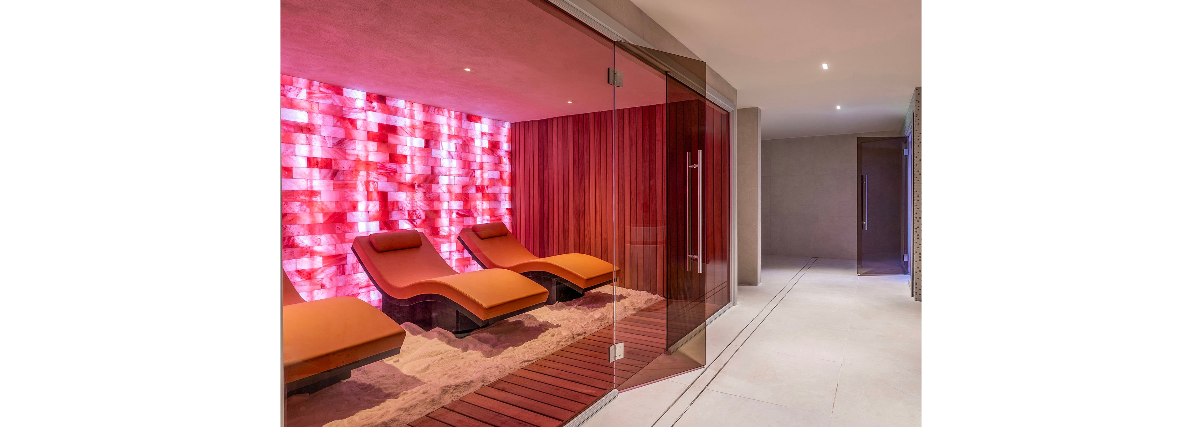A contemporary spa relaxation room features several orange reclining loungers arranged on a sand-covered floor. The space is enclosed by glass walls, revealing warm wooden paneling and a striking illuminated wall with pink and red tones. Clean, minimalist corridors extend beyond the room, emphasizing a serene and luxurious atmosphere. Soft lighting and sleek surfaces create a calm, modern wellness environment.