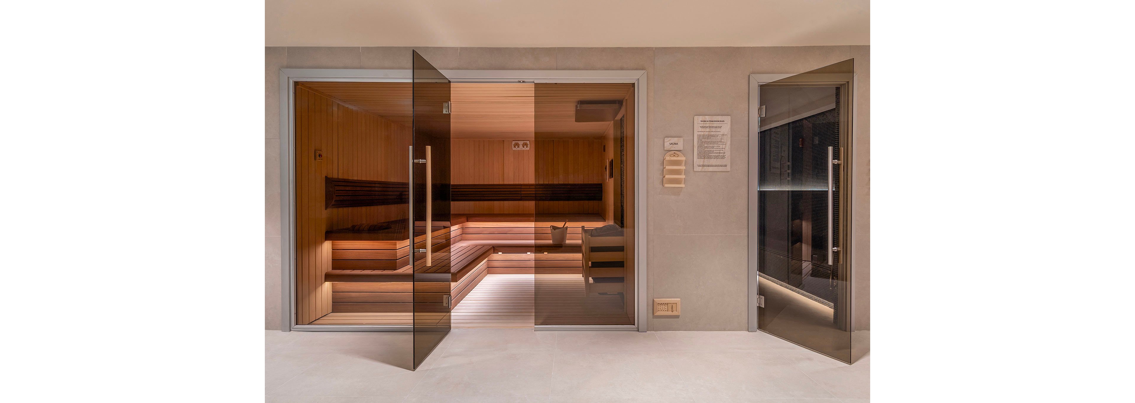 A contemporary wooden sauna room is viewed through large glass doors in a spa or wellness facility. Warm interior lighting highlights the tiered wooden benches and clean minimalist design. Adjacent to the sauna, another glass door leads to a separate room with darker lighting. Light-colored tile flooring and neutral walls create a calm, luxurious atmosphere.