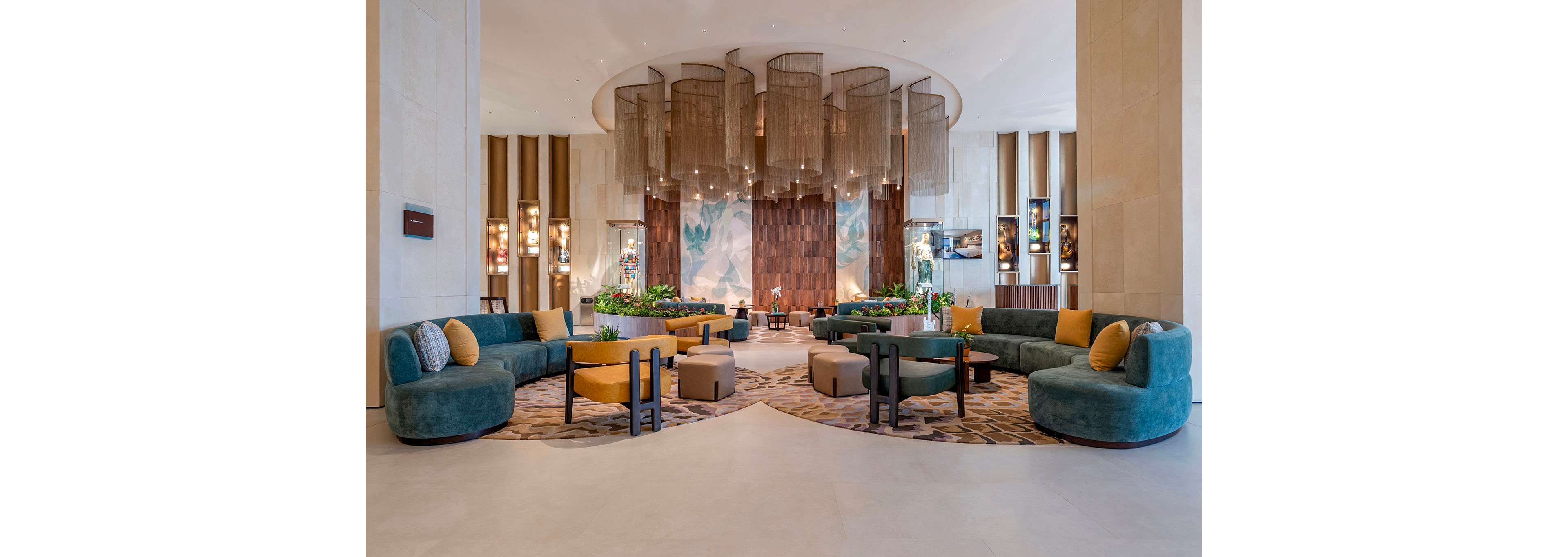 A spacious contemporary lobby features curved teal sofas and mustard accent cushions arranged symmetrically around low wooden tables. Tall sculptural metal screens hang from the ceiling, framing a textured wood and abstract mural backdrop. Warm lighting, indoor plants, and patterned carpeting create a welcoming, upscale atmosphere. The overall design blends modern luxury with soft, earthy tones and organic shapes.