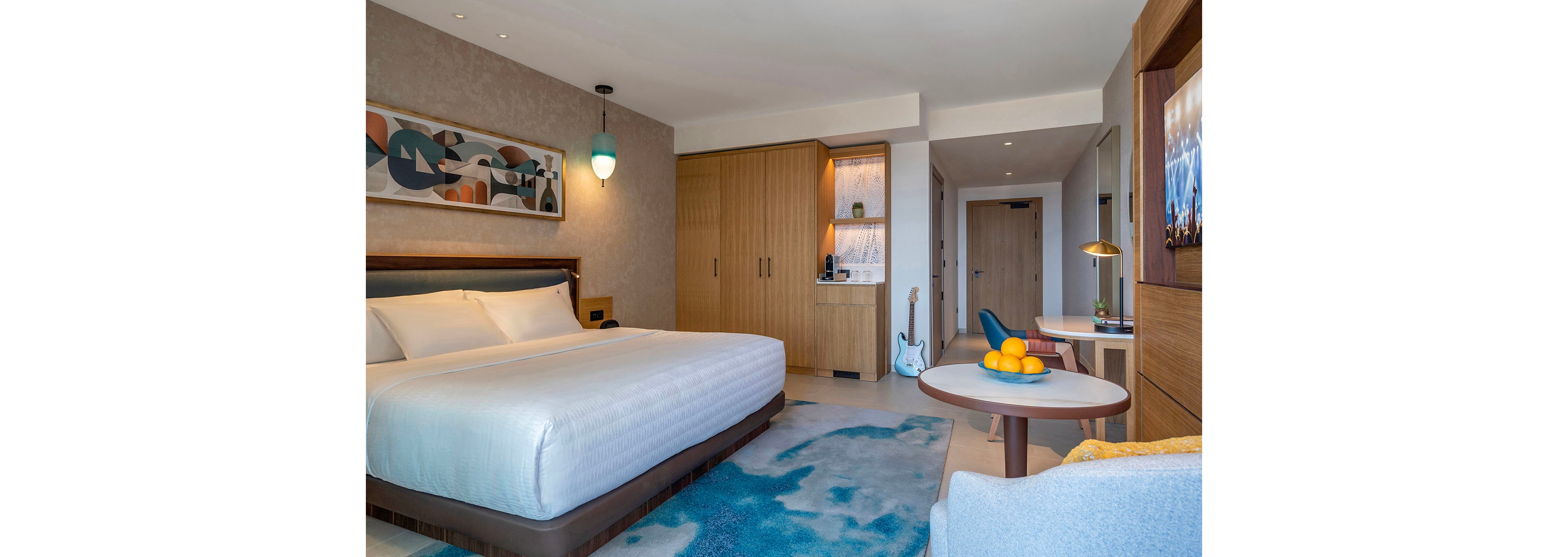 A contemporary hotel room features a large king-size bed with crisp white linens and a padded headboard. Built-in wooden wardrobes and a small kitchenette area with shelving sit along the back wall, while a guitar rests near the doorway. A round table with a bowl of oranges and a light blue armchair add color beside a soft blue area rug. Abstract wall art and warm lighting create a relaxed, inviting atmosphere for guests.