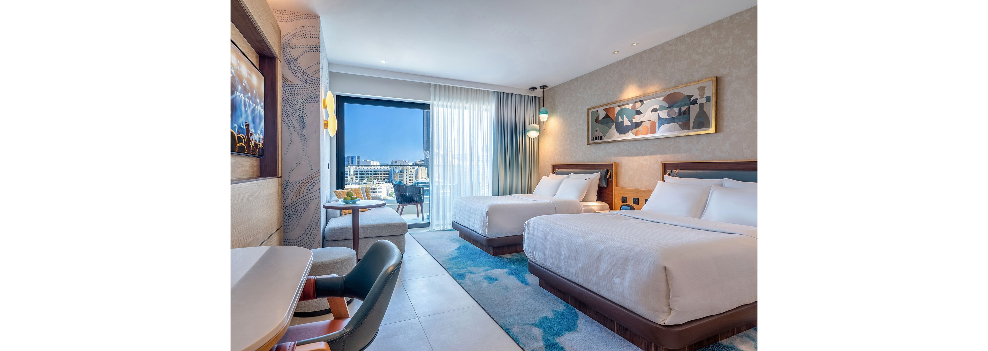 A contemporary hotel room features two large beds with crisp white linens and a soft blue area rug. Floor-to-ceiling windows with sheer curtains open to a balcony overlooking a cityscape and distant buildings. A small table with chairs sits near the window, while a desk and seating area occupy the foreground. Warm accent lighting and abstract wall art create a relaxed, upscale atmosphere.