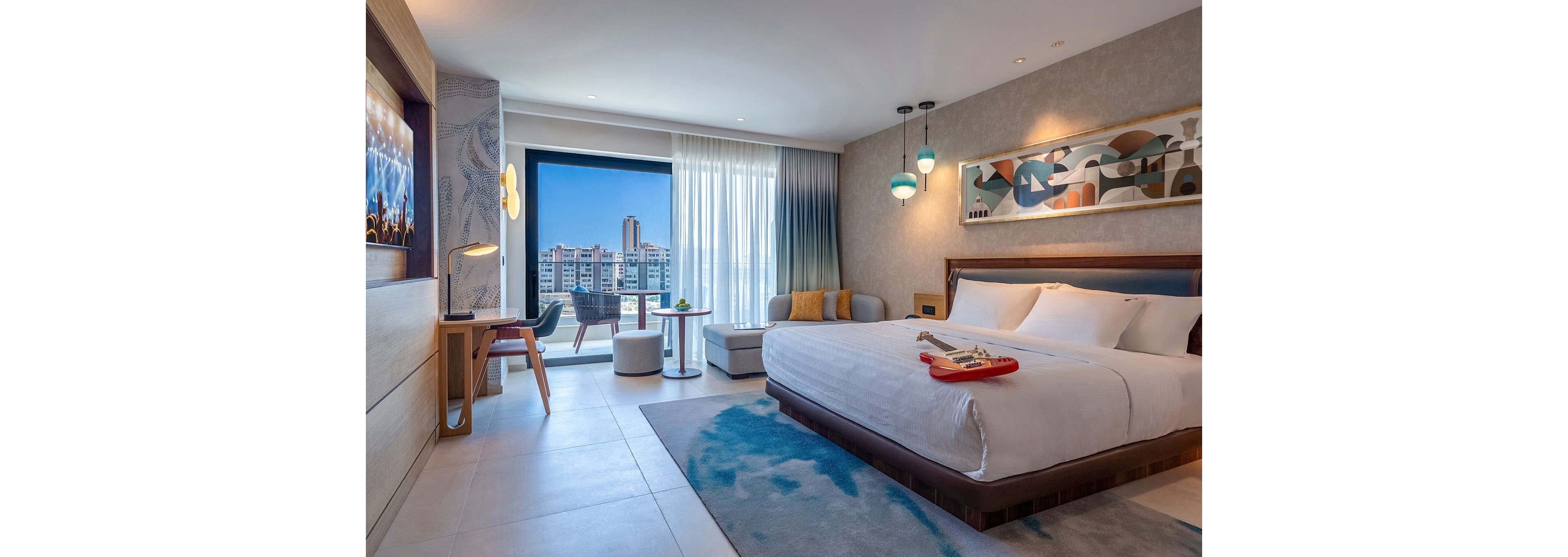 Contemporary hotel bedroom featuring a large double bed and stylish decor. A floor-to-ceiling window reveals a bright city skyline and blue sky outside. The room includes a desk area, cozy seating corner, abstract wall art, and colorful pendant lights. Soft neutral tones with blue accents create a clean, inviting atmosphere.