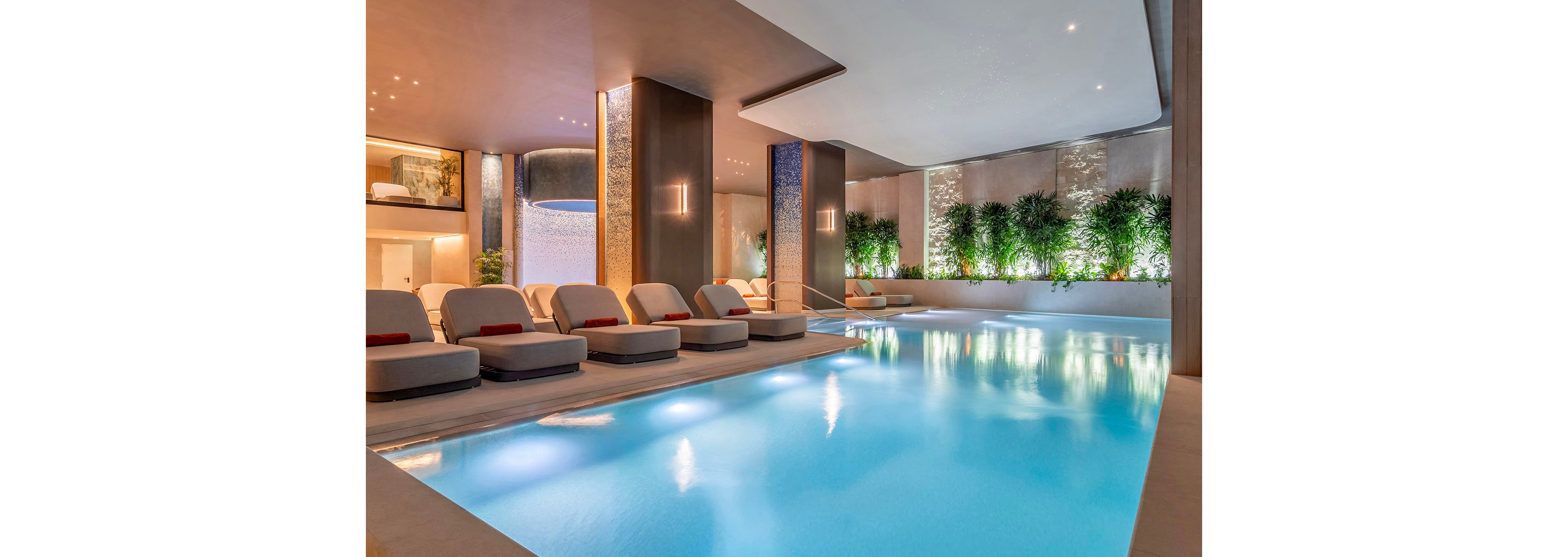 A serene indoor spa pool is surrounded by modern lounge chairs and soft ambient lighting. Tall columns and textured walls frame the water, creating a sophisticated architectural setting. A row of lush green plants lines one side of the pool, adding a natural touch to the contemporary design. The overall atmosphere is calm, inviting, and designed for relaxation and wellness.