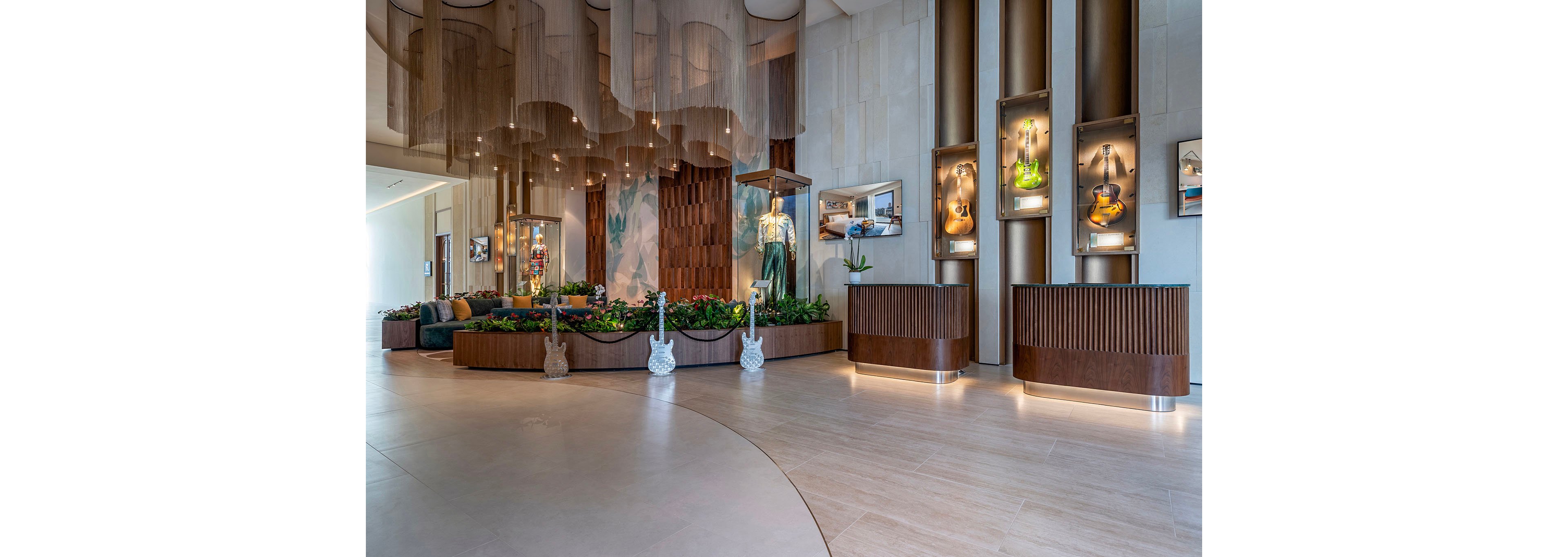 A spacious contemporary lobby features sculptural lighting and a central planter filled with lush greenery. Framed guitars are displayed vertically on the wall, adding a musical and artistic focal point to the space. Warm wood tones, soft seating in the background, and hanging light fixtures create an inviting atmosphere. The clean, light-colored floor and high ceiling emphasize openness and modern design.
