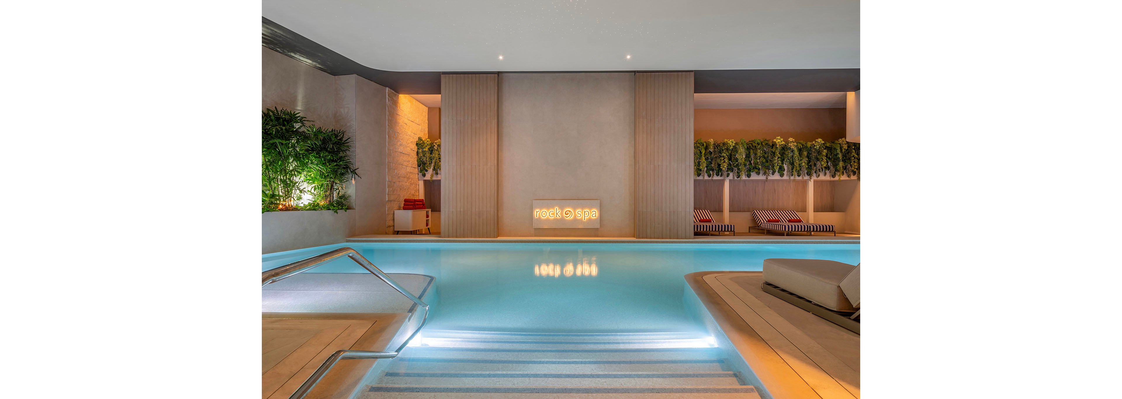 A serene indoor spa pool is illuminated with soft, ambient lighting and framed by minimalist architectural details. Steps lead gently into the turquoise water, with a metal handrail on the left side. Green plants and hanging foliage add a natural touch above cushioned lounge beds with striped covers. A softly lit sign reading "rock spa" appears on the far wall, enhancing the relaxing, contemporary atmosphere.