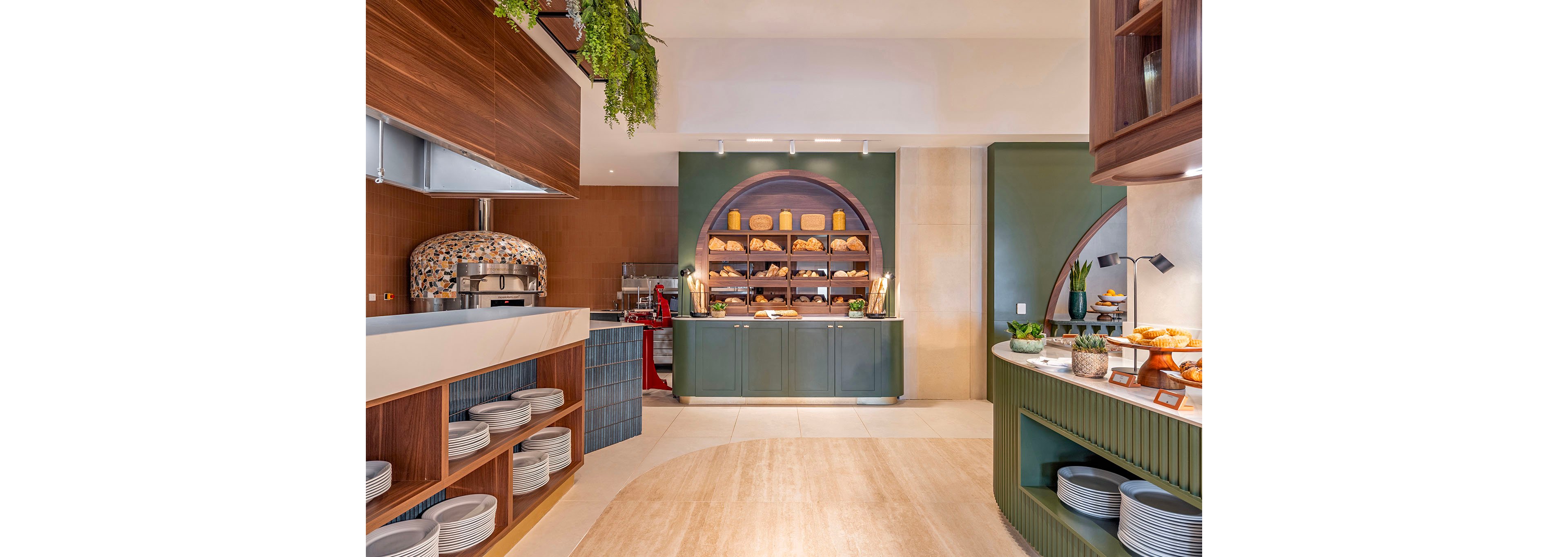 A contemporary bakery buffet area showcases shelves filled with assorted loaves of bread and pastries. The setting features warm wood accents, green cabinetry, and a tiled pizza oven in the background. Plates are neatly stacked on open shelving, and a side counter presents fruit, croissants, and other breakfast items. Soft lighting and natural tones create an inviting, upscale dining atmosphere.