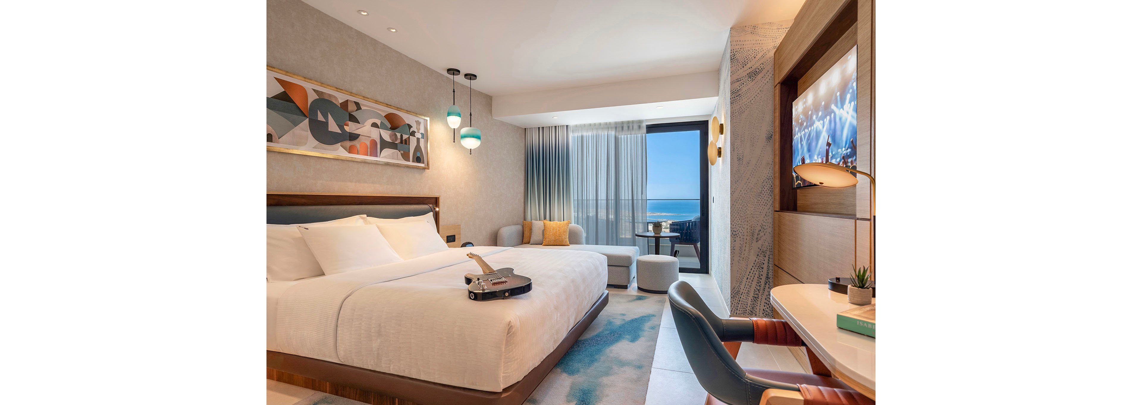 A contemporary hotel room features a large bed with crisp white linens and a guitar resting on top. The space includes stylish pendant lights, abstract wall art, and a cozy seating area near floor-to-ceiling windows. Sheer curtains frame a balcony that overlooks a bright blue ocean horizon. A desk with a chair, books, and a television displaying a concert scene complete the modern, relaxing atmosphere.