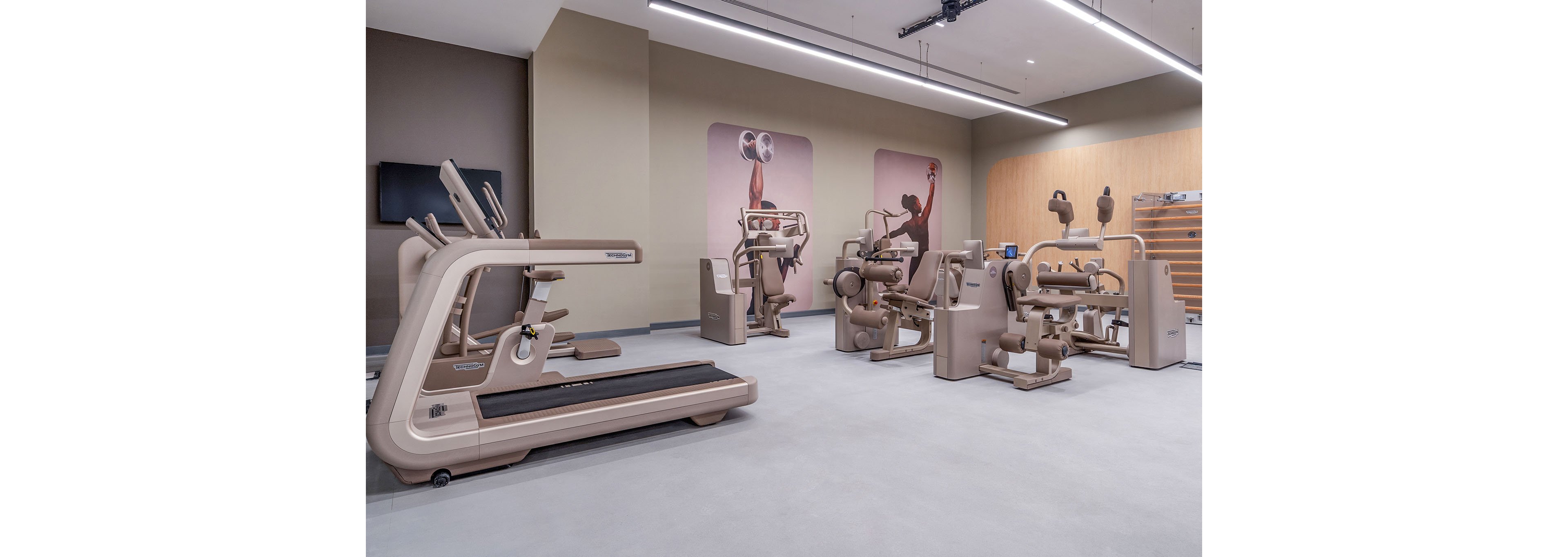 A contemporary gym interior features a collection of beige exercise machines, including a treadmill and various strength-training equipment. The space is clean and spacious with light flooring and neutral walls. Large wall graphics show silhouettes of people working out, adding a dynamic visual element. Overhead linear lighting and a mounted screen contribute to the modern, professional atmosphere.