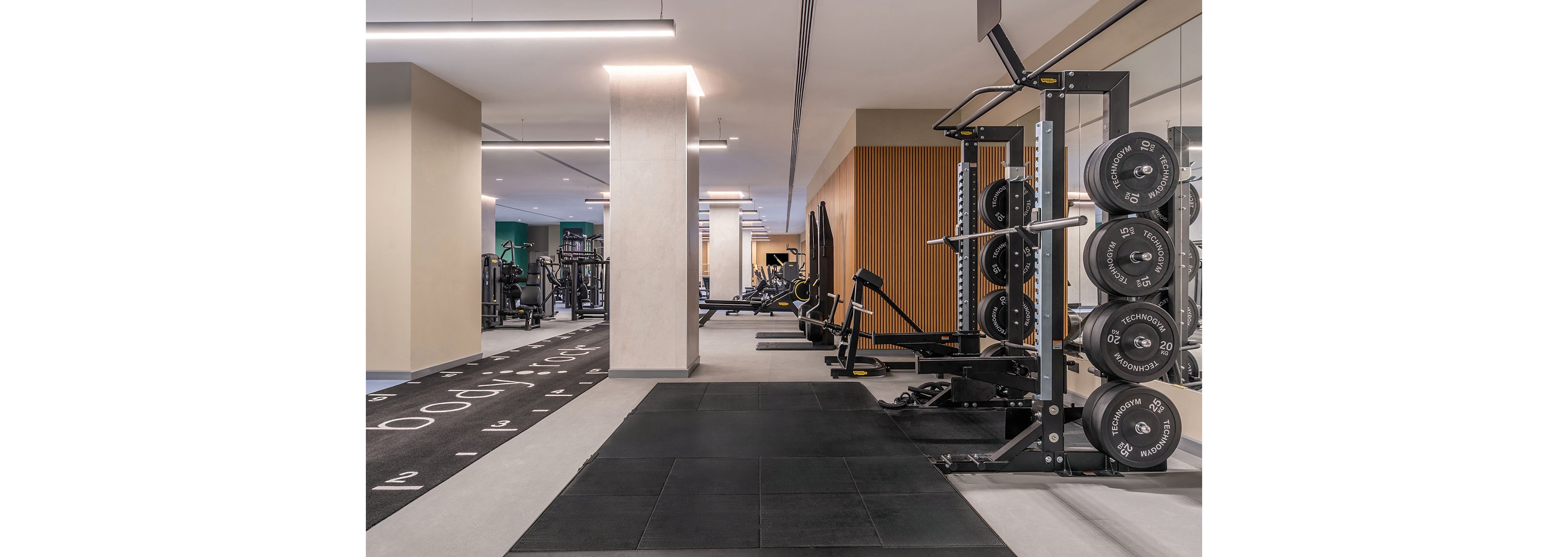 A contemporary fitness center interior features a spacious weightlifting area with a squat rack and neatly stacked weight plates. The room includes rubber flooring, a functional training track, and various strength machines in the background. Bright, even lighting and neutral walls create a clean, minimalist atmosphere. No people are present, emphasizing the equipment and layout of the gym space.