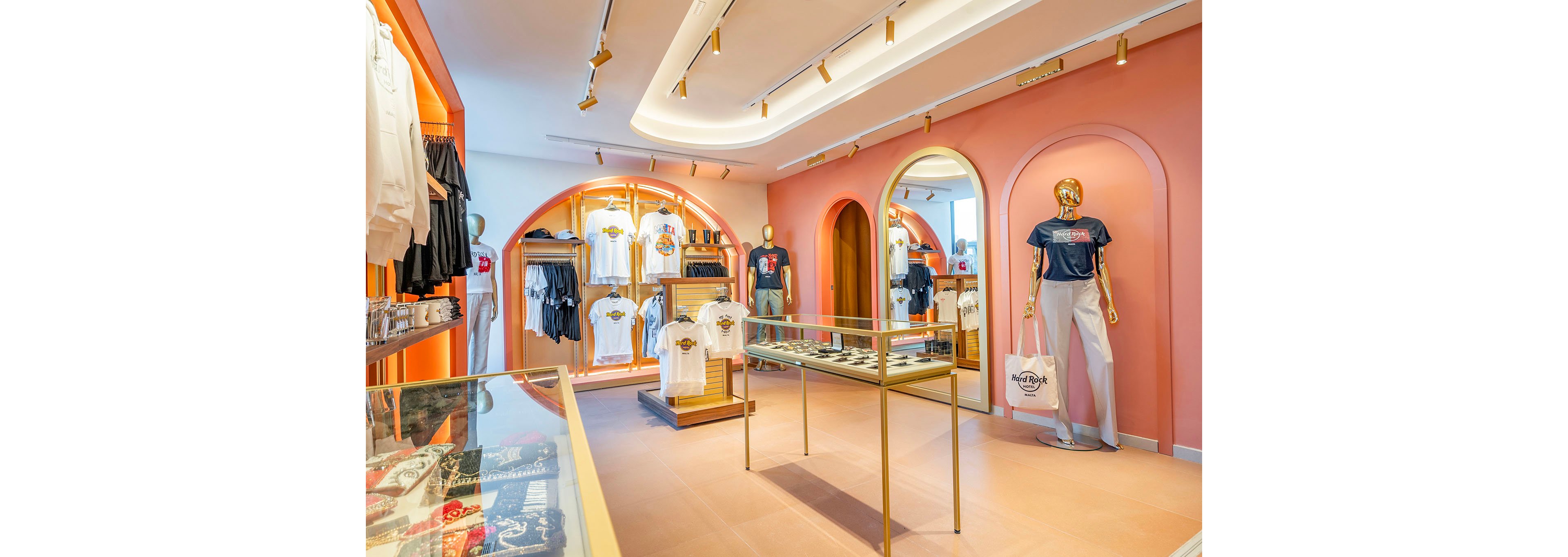 Contemporary retail boutique featuring neatly arranged t-shirts and apparel on wooden racks. The setting includes arched wall niches, glass display cases, and warm ambient lighting against peach-colored walls. A gold mannequin stands near the entrance holding a white shopping bag, adding a stylish focal point. Overall, the scene conveys a clean, fashionable, and inviting shopping environment.