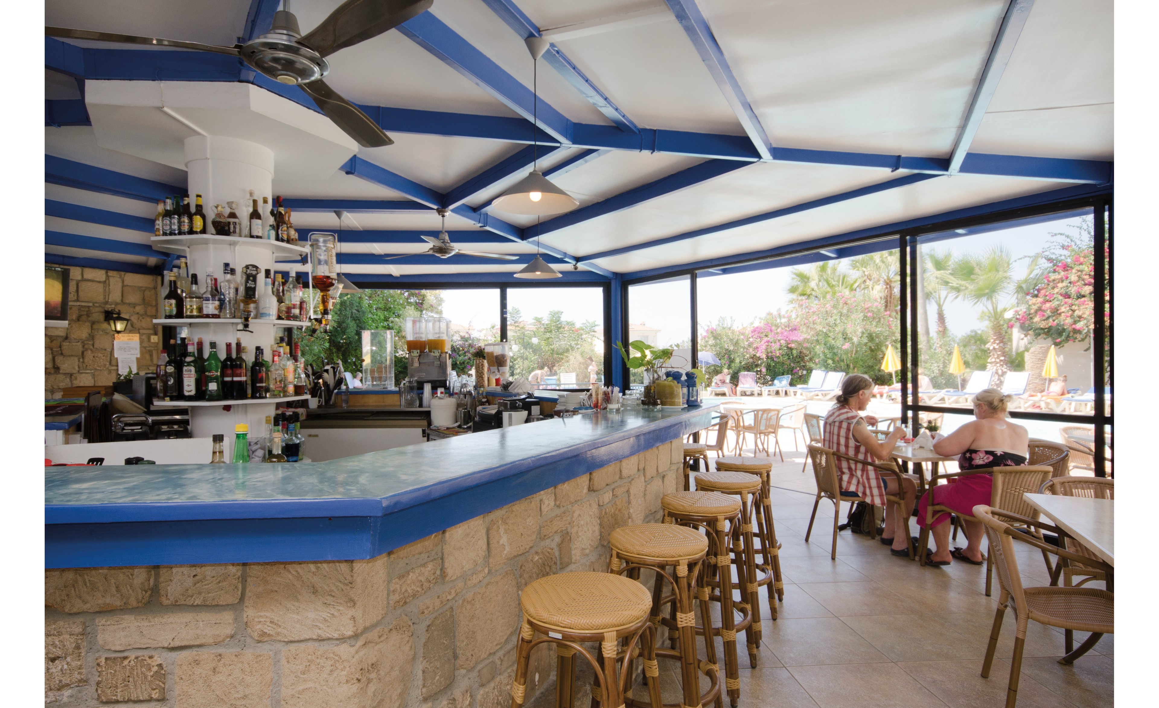 An open-air bar with a stone counter and blue accents, featuring a variety of bottles and glasses neatly arranged. The setting includes wicker chairs and tables, with two women seated enjoying the tropical ambiance. Large windows provide a view of palm trees, vibrant flowers, and sunny weather.