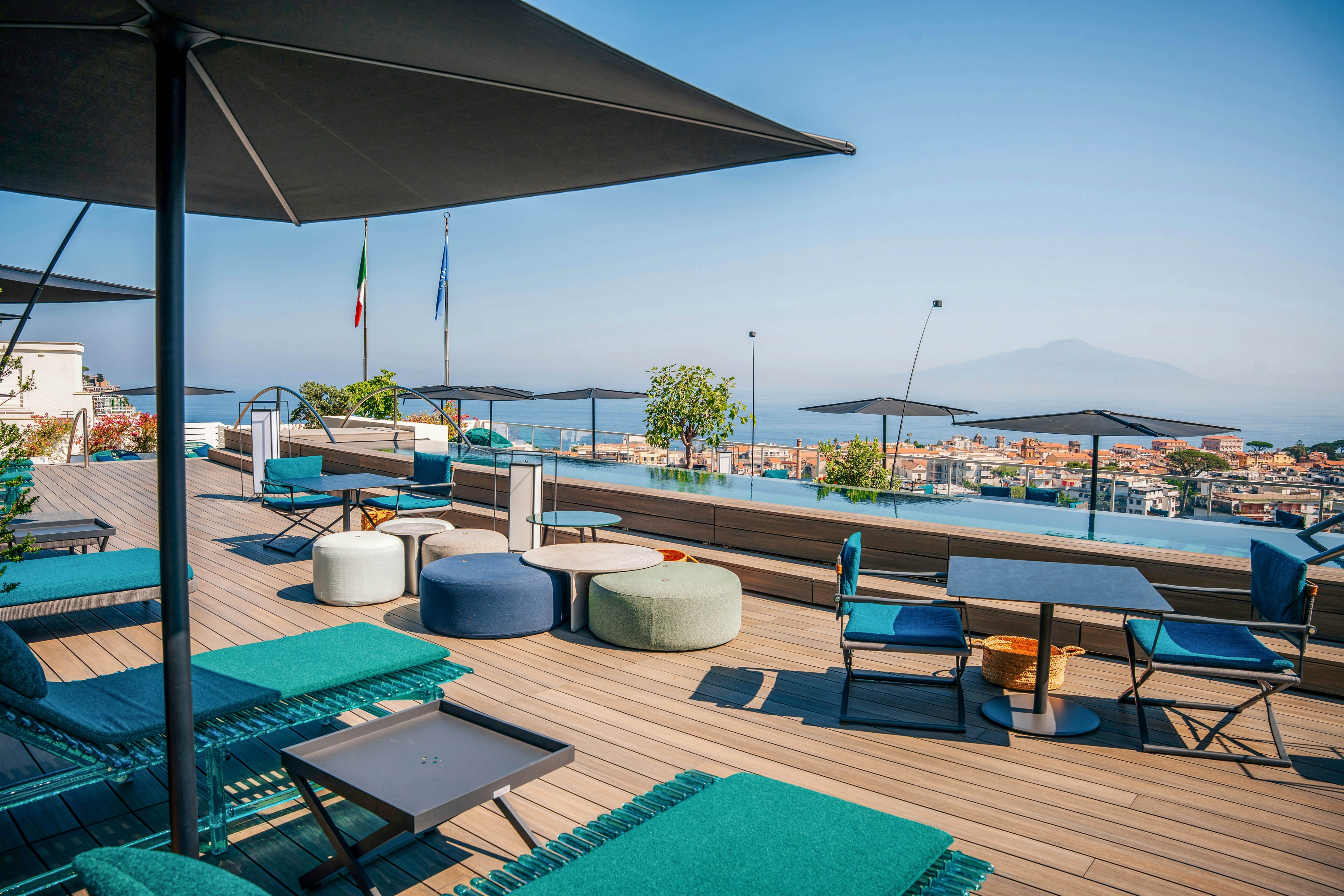 A stylish rooftop terrace features contemporary outdoor seating, tables, and umbrellas overlooking a cityscape and distant mountains. The setting includes a sleek infinity pool and visible flags, suggesting a cosmopolitan atmosphere. The color palette is dominated by cool blues and neutrals, creating a relaxed and inviting mood. No people are present, emphasizing tranquility and leisure.