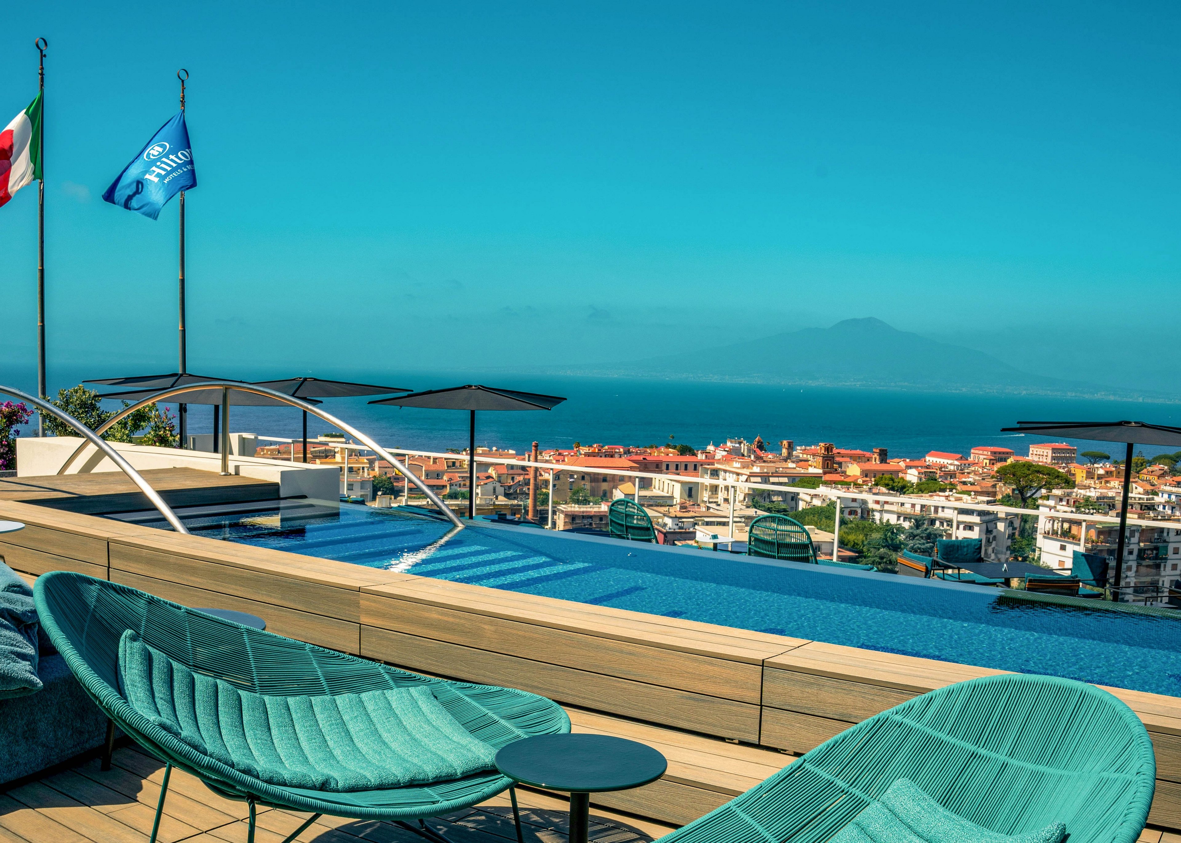 A stylish rooftop pool area features turquoise lounge chairs and a wooden deck, set against a panoramic view of a coastal city and distant mountains. Two flags, including one with visible text, wave in the breeze above the pool. The scene is bathed in bright daylight, with clear blue skies and a tranquil atmosphere. The overall mood is relaxed and luxurious, ideal for leisure and vacation settings.