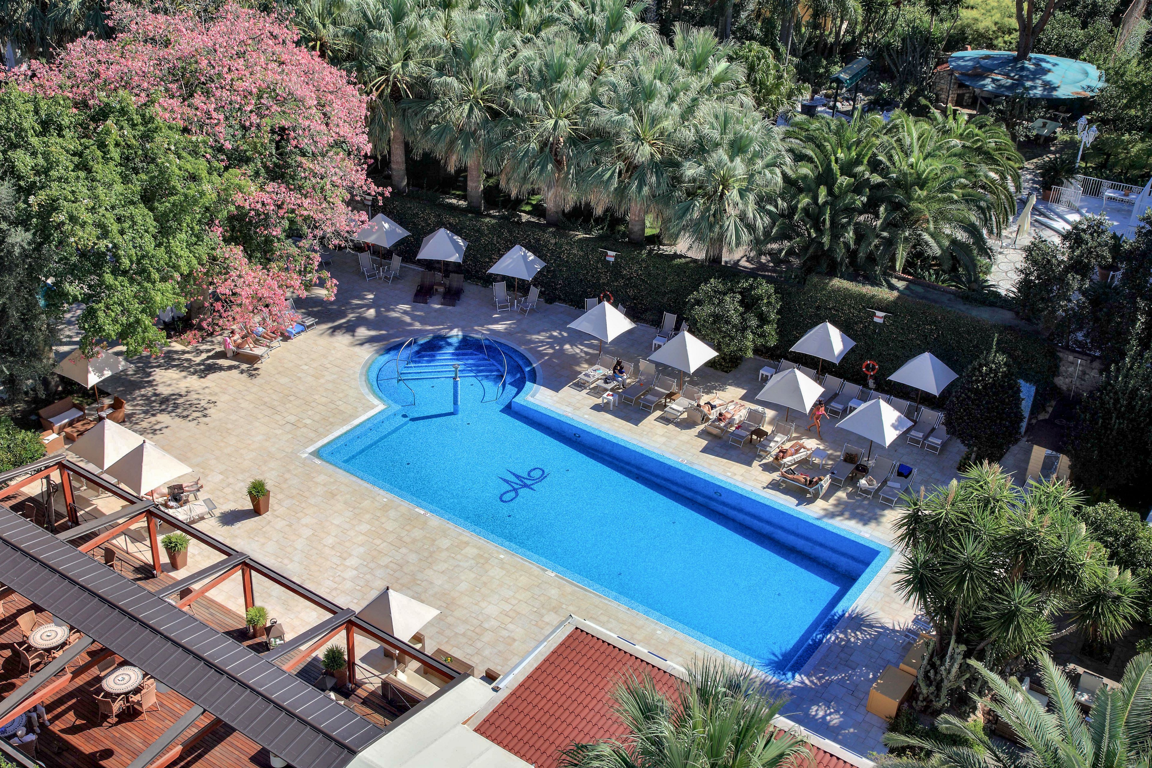 A large rectangular swimming pool is seen from above in a lush resort setting. Sun loungers and white umbrellas line the poolside terrace, with guests relaxing around the water. A smaller circular pool adjoins the main pool, and the blue pool floor features stylized text at the center. Surrounding palm trees and flowering greenery create a tropical, tranquil atmosphere.