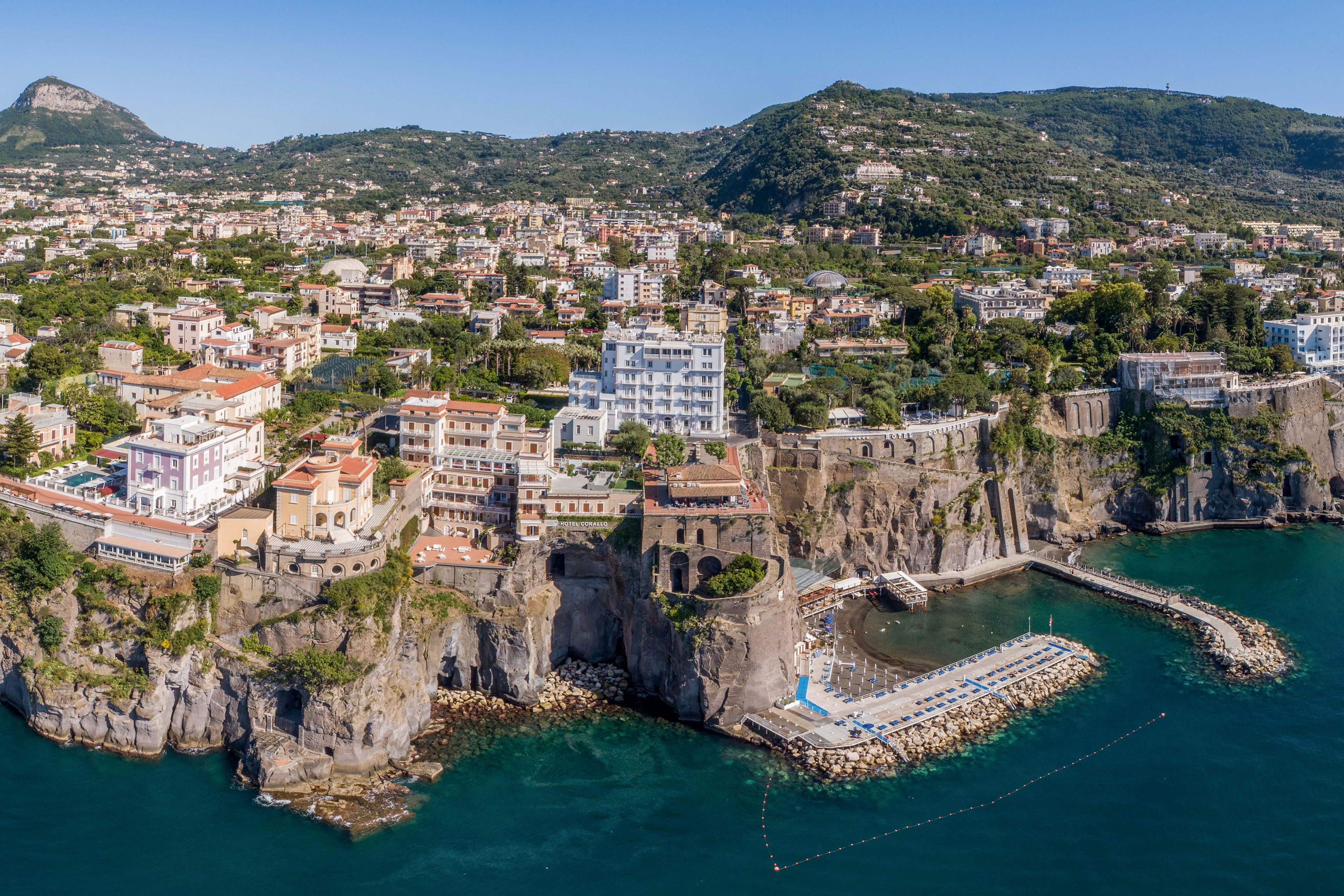 A coastal town stretches along steep cliffs overlooking a calm blue sea. Colorful buildings and hotels line the cliff edge, with terraced structures descending toward the waterfront. A small marina and beach area with sun loungers and umbrellas extend into the water. Green hills rise in the background, framing the densely built seaside community under a clear sky.