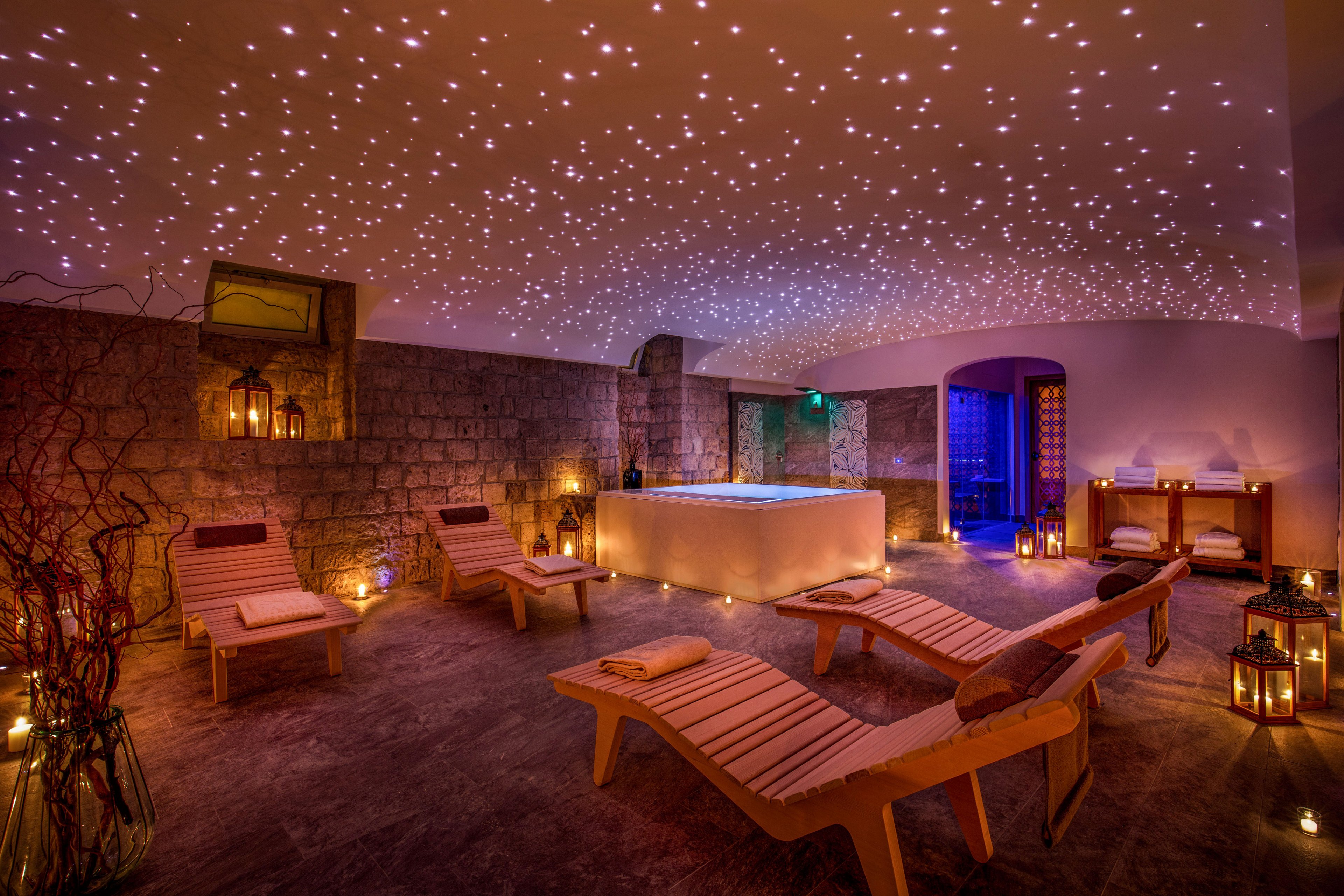 A tranquil spa lounge features several reclining chairs arranged around a central illuminated hot tub. Warm candlelight and lanterns create a cozy atmosphere against stone walls and soft flooring. The ceiling is covered with tiny star-like lights, adding a magical, relaxing ambiance. Wooden furniture, decorative branches, and subtle colored lighting in the background enhance the sense of luxury and calm.