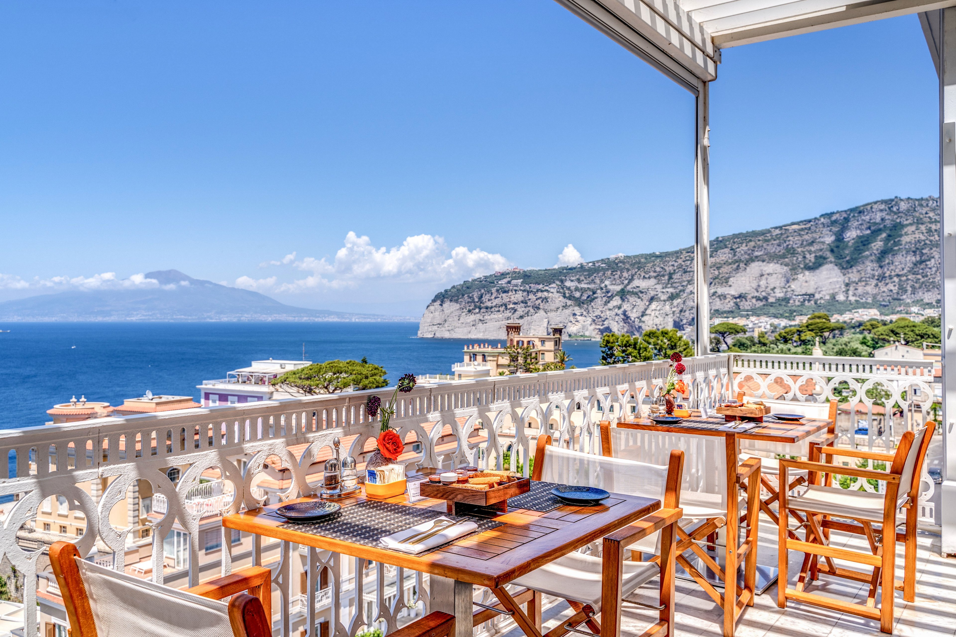 An open-air restaurant terrace overlooks a bright blue sea and distant mountains under a clear sky. Wooden tables and chairs are neatly arranged with place settings, baskets, and decorative items. Ornate white railings frame the balcony, adding elegance to the coastal setting. The scene feels calm and inviting, suggesting a leisurely meal with panoramic waterfront views.