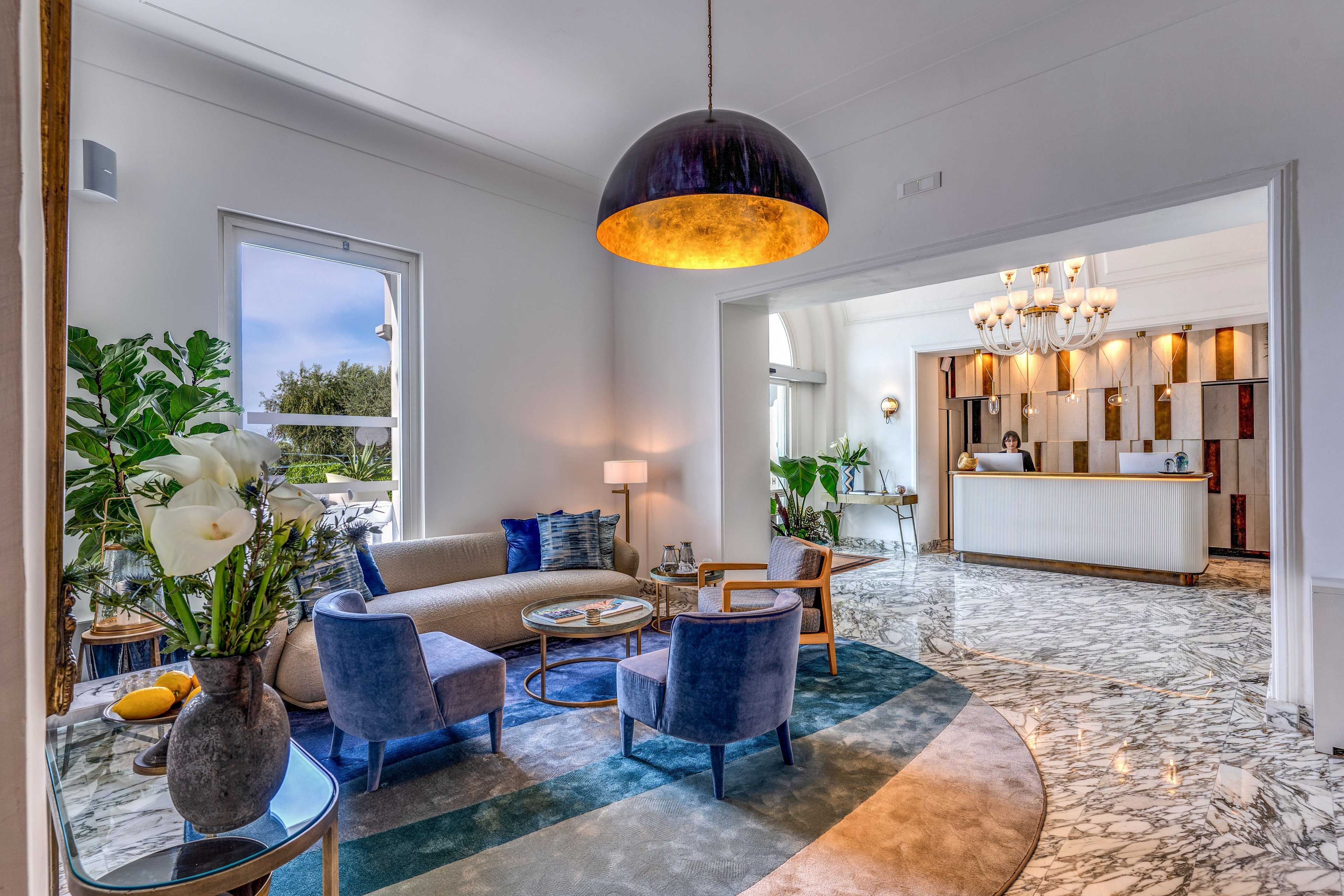 A stylish lounge area features a curved sofa and blue velvet armchairs arranged around small coffee tables. Large windows bring in natural light and offer a view of greenery outside. A dramatic dome pendant light and a crystal chandelier illuminate the marble floor and modern decor. Potted plants, decorative cushions, and fresh flowers add color and texture to the sophisticated interior.