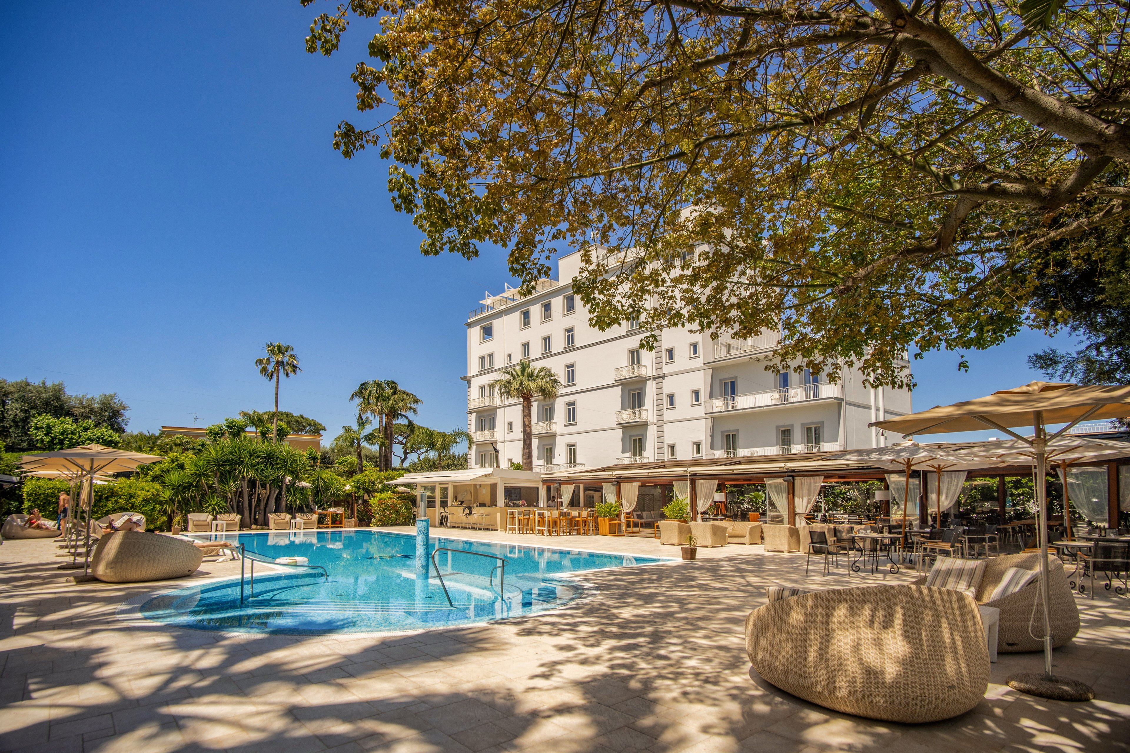 An outdoor swimming pool sits at the center of a sunny hotel courtyard. Lounge chairs, umbrellas, and shaded seating areas surround the pool, creating a relaxed vacation atmosphere. A multi-story white hotel building rises in the background, framed by palm trees and a large leafy tree. The clear blue water and bright daylight suggest a warm, inviting resort setting ideal for leisure and relaxation.