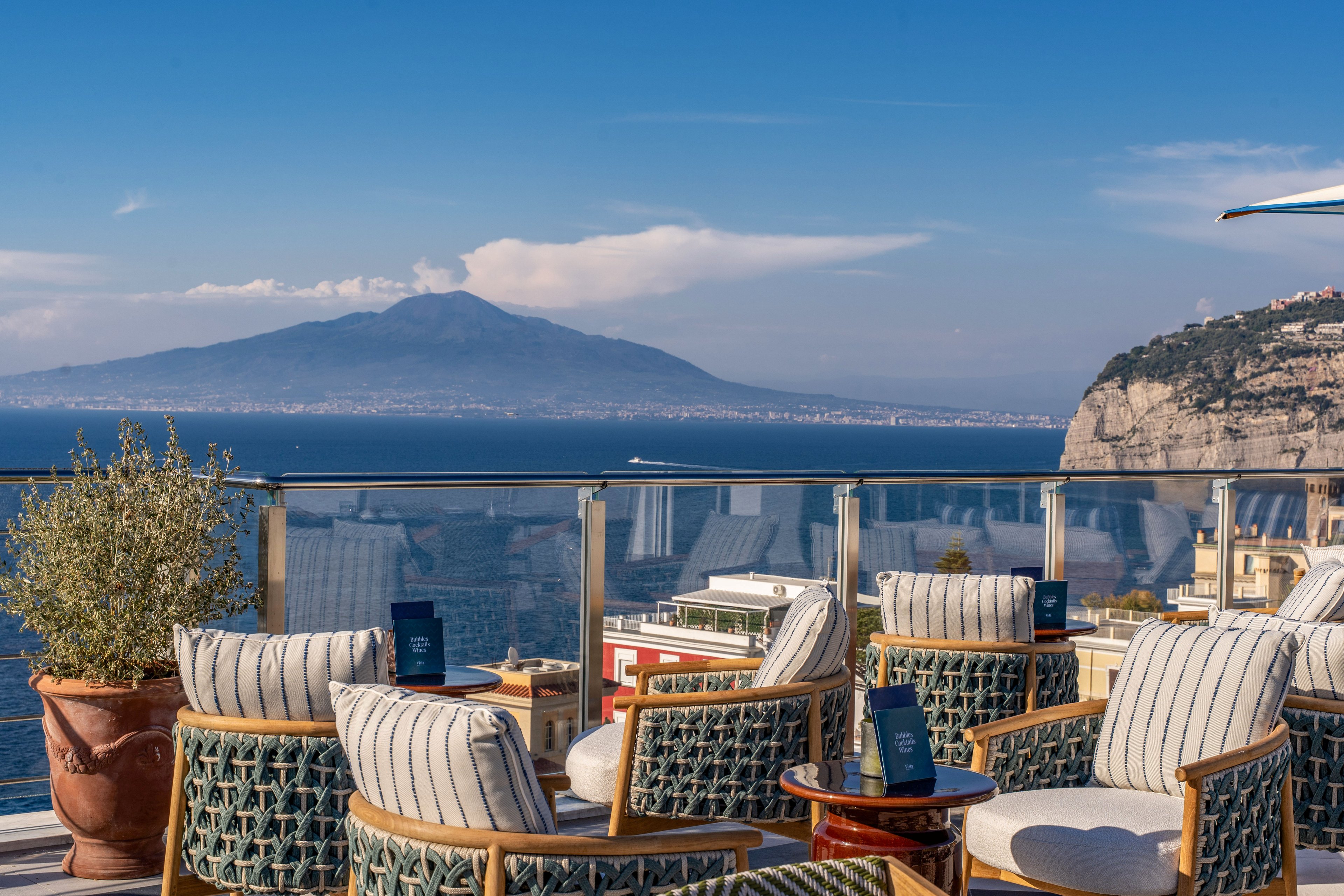An elegant rooftop terrace features cushioned wicker chairs arranged around small tables with menus. Glass railings frame a sweeping view of the sea and a distant mountain under a clear blue sky. A potted plant and striped upholstery add texture and comfort to the outdoor seating area. The scene suggests a relaxed, upscale setting ideal for leisure, dining, or enjoying the panoramic coastal scenery.