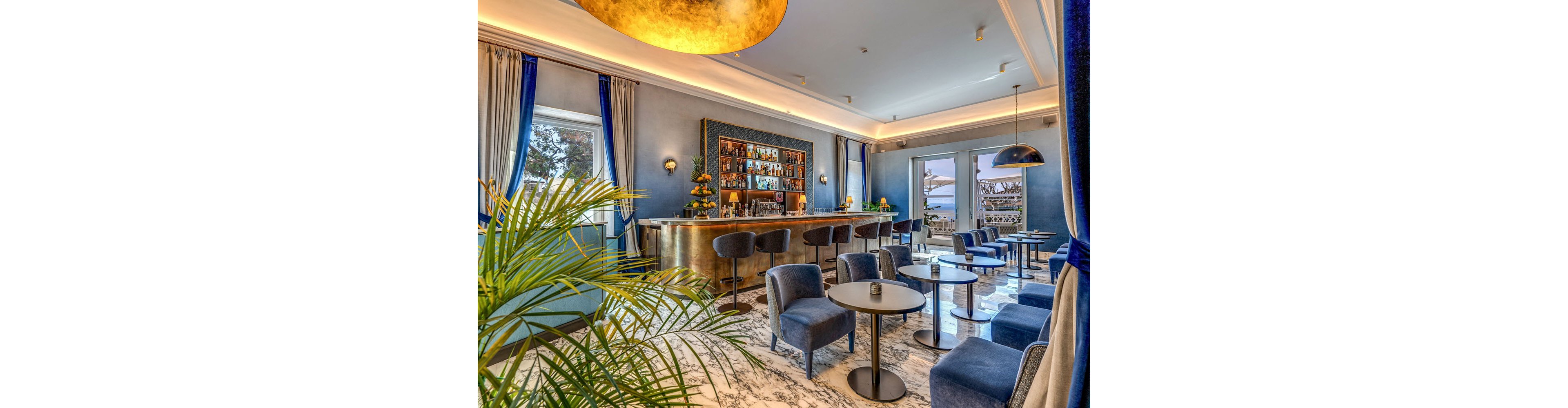 Stylish lounge bar interior featuring a golden counter and plush blue seating. The space is decorated with marble flooring, round tables, and contemporary pendant lighting. Large windows and glass doors open to a terrace with a scenic ocean view, bringing natural light into the room. Green plants and blue curtains add a fresh, sophisticated touch to the modern design.
