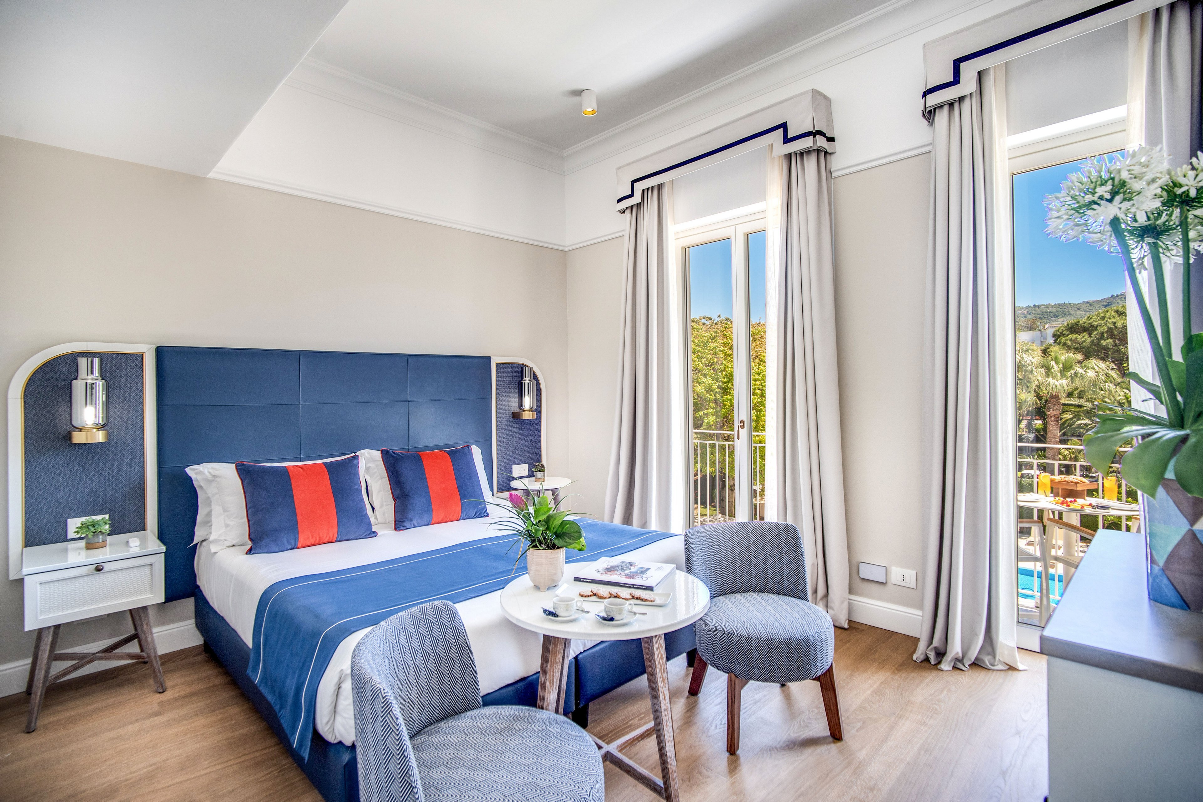 Contemporary hotel bedroom features a double bed with blue headboard and colorful striped pillows. A small round table with chairs and potted plants sits on wooden flooring, adding a cozy seating area. Large windows with light curtains open to a balcony overlooking greenery, palm trees, and a pool. The room is decorated in a fresh blue and white palette with natural light enhancing the airy atmosphere.