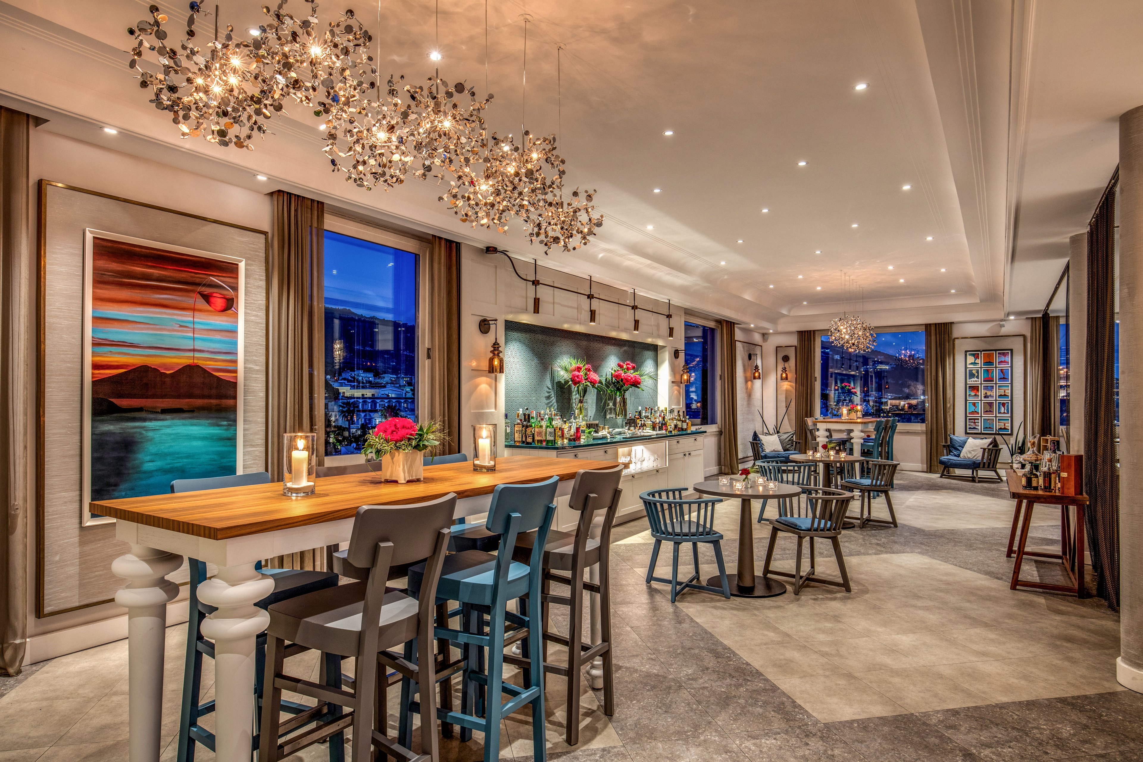 A stylish hotel bar lounge features high tables and modern chairs arranged in a spacious interior. Large windows reveal a vibrant nighttime cityscape with twinkling lights outside. Decorative chandeliers, colorful artwork, and floral arrangements add sophistication and warmth to the space. The bar counter is neatly stocked with bottles and glassware, creating an inviting atmosphere for evening socializing.