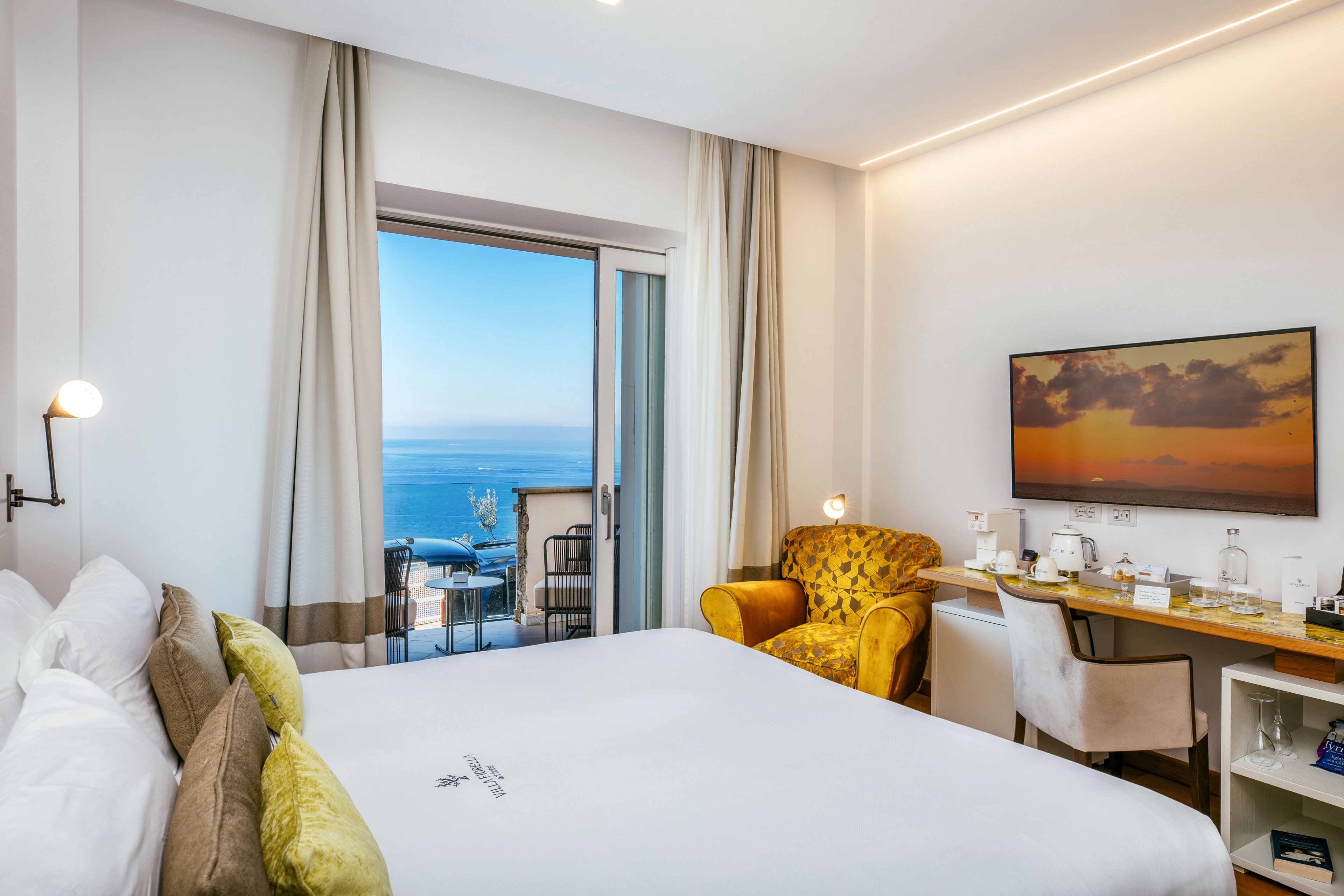 Bright contemporary hotel room features a large bed facing a balcony with a clear ocean view. A glass door and tall curtains frame the outdoor seating area overlooking the sea. Inside, a yellow armchair, wall-mounted television, and neatly arranged desk with refreshments create a comfortable atmosphere. Soft lighting and neutral tones give the space a calm, inviting feel.