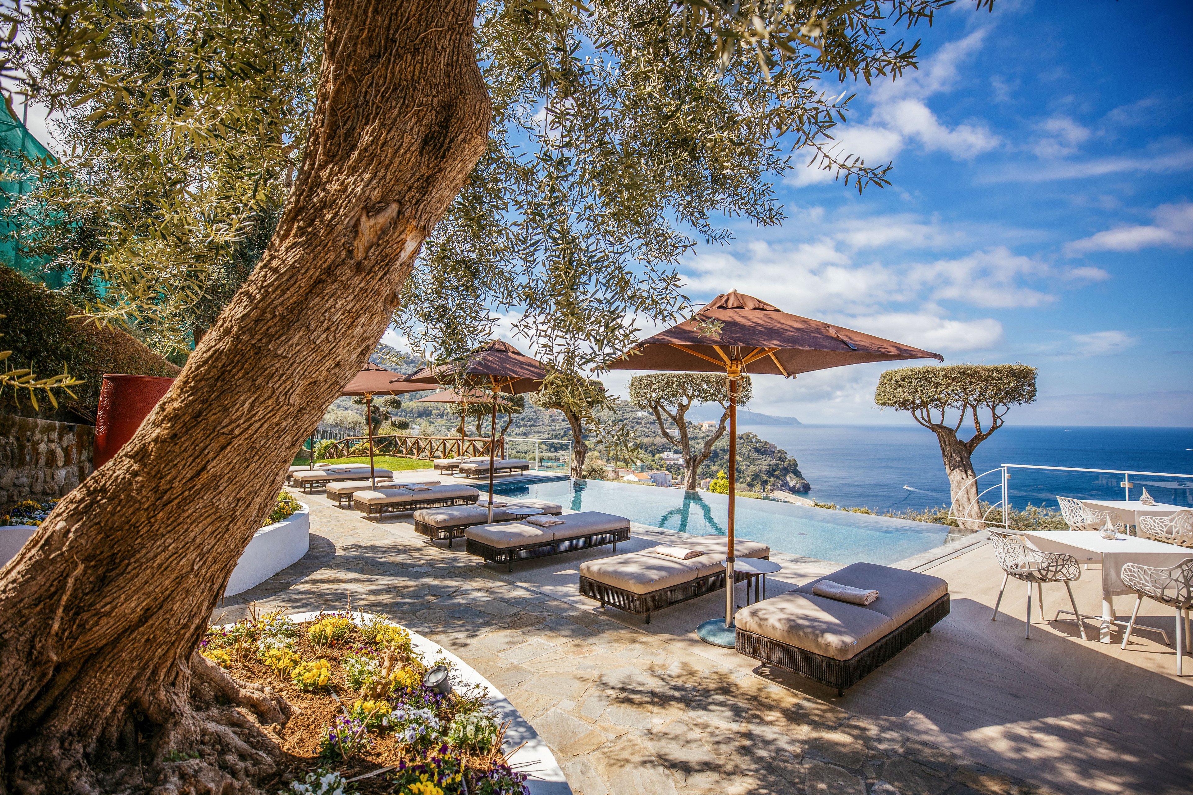 A sunlit terrace features an infinity pool overlooking a bright blue sea and rugged coastline. Cushioned sun loungers and wooden umbrellas line the poolside, creating a relaxing resort atmosphere. Sculpted trees, flowering plants, and outdoor café chairs add decorative detail to the scene. Clear skies with scattered clouds enhance the warm, tranquil Mediterranean-style setting.