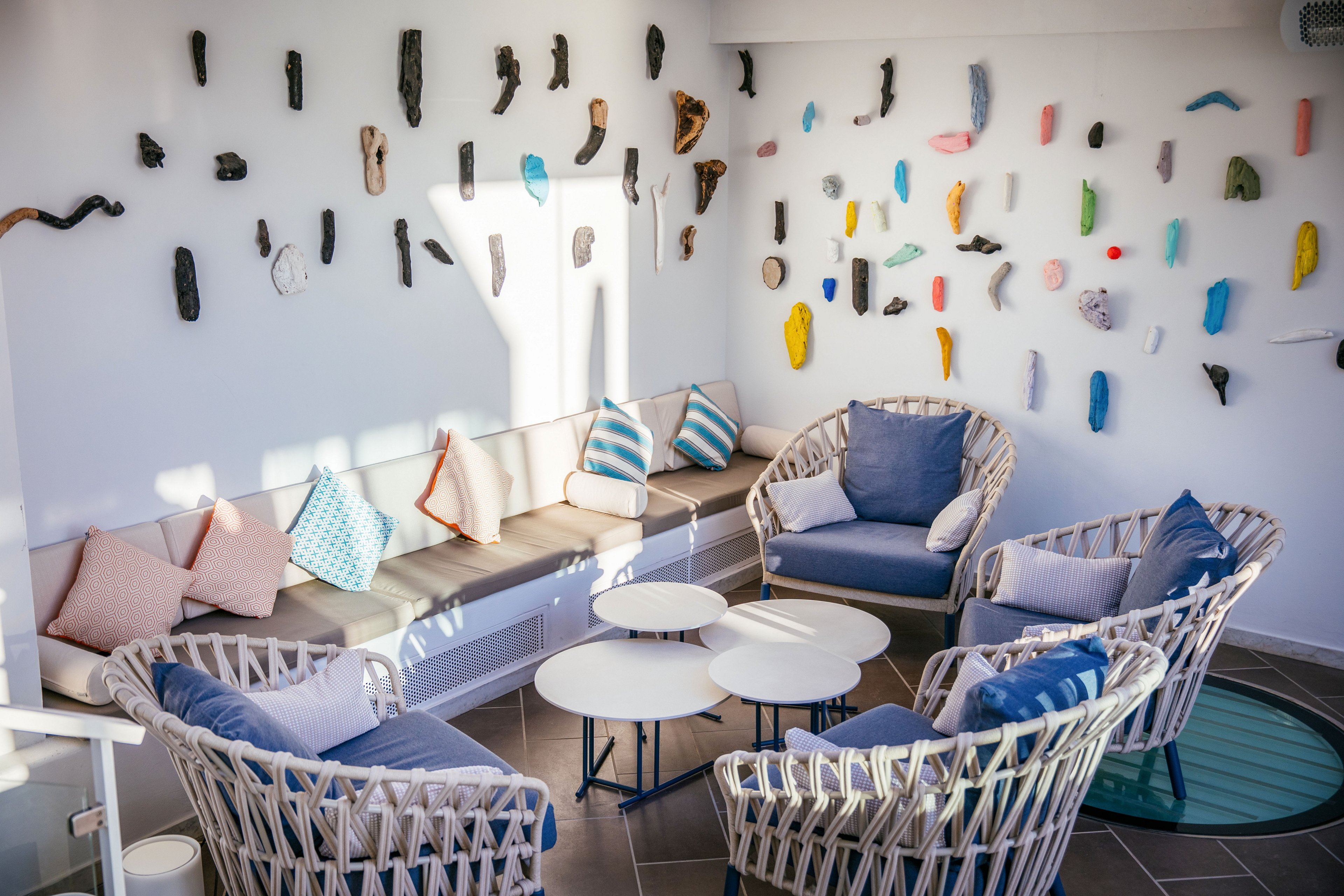 Contemporary lounge seating is arranged around small round tables in a bright, sunlit room. Woven armchairs and cushioned bench seating feature pastel and blue-toned pillows, creating a relaxed coastal atmosphere. The white wall is decorated with numerous abstract sculptural pieces in various shapes and colors. Light streaming in enhances the airy, minimalist design and highlights the playful art installation.