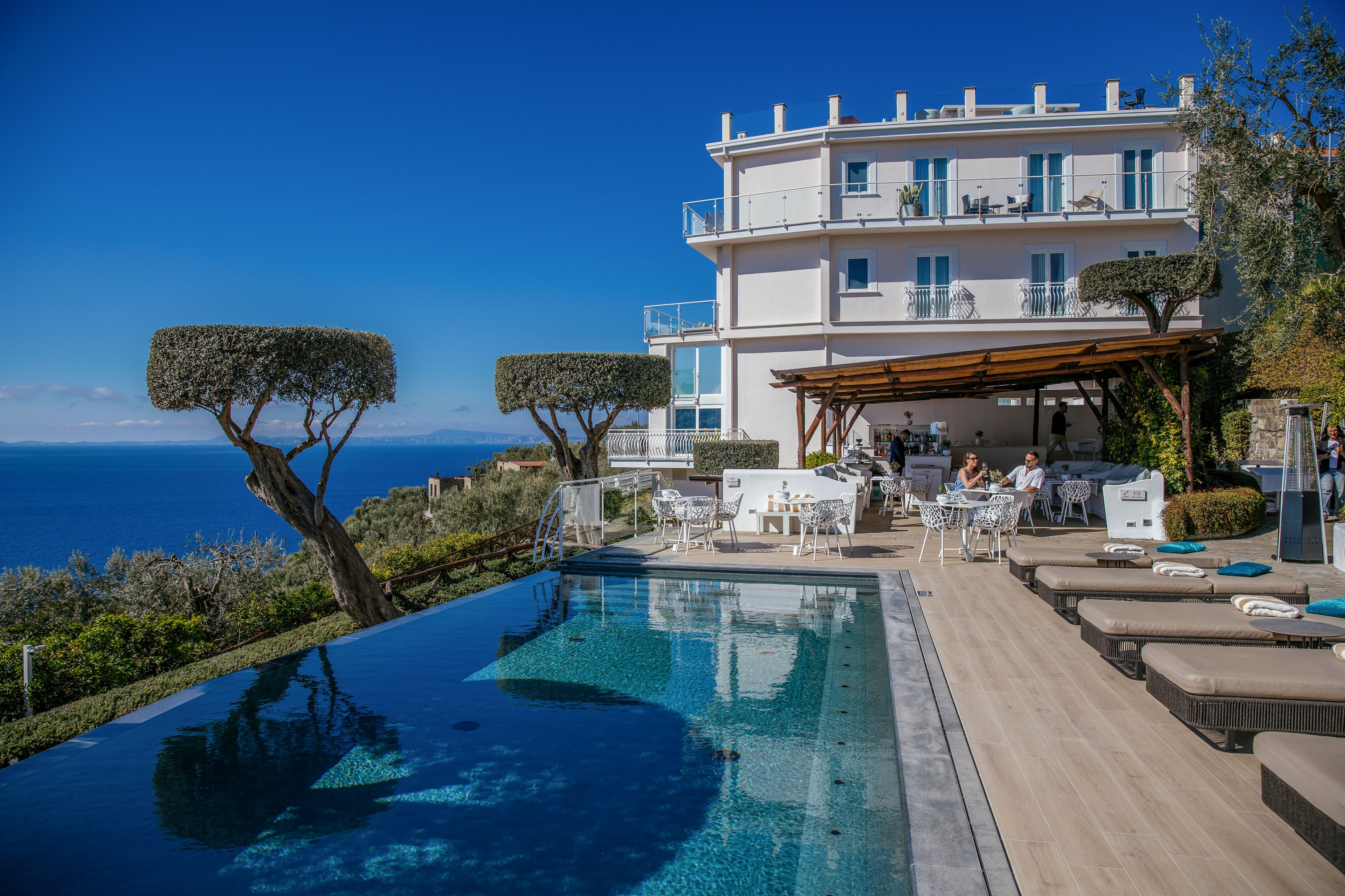 A modern seaside hotel overlooks a deep blue ocean from an elevated terrace. The foreground features an infinity pool beside cushioned sun loungers and manicured trees. Guests sit at white outdoor tables under a wooden pergola attached to the building. Clear skies and bright sunlight create a relaxed, upscale vacation atmosphere.